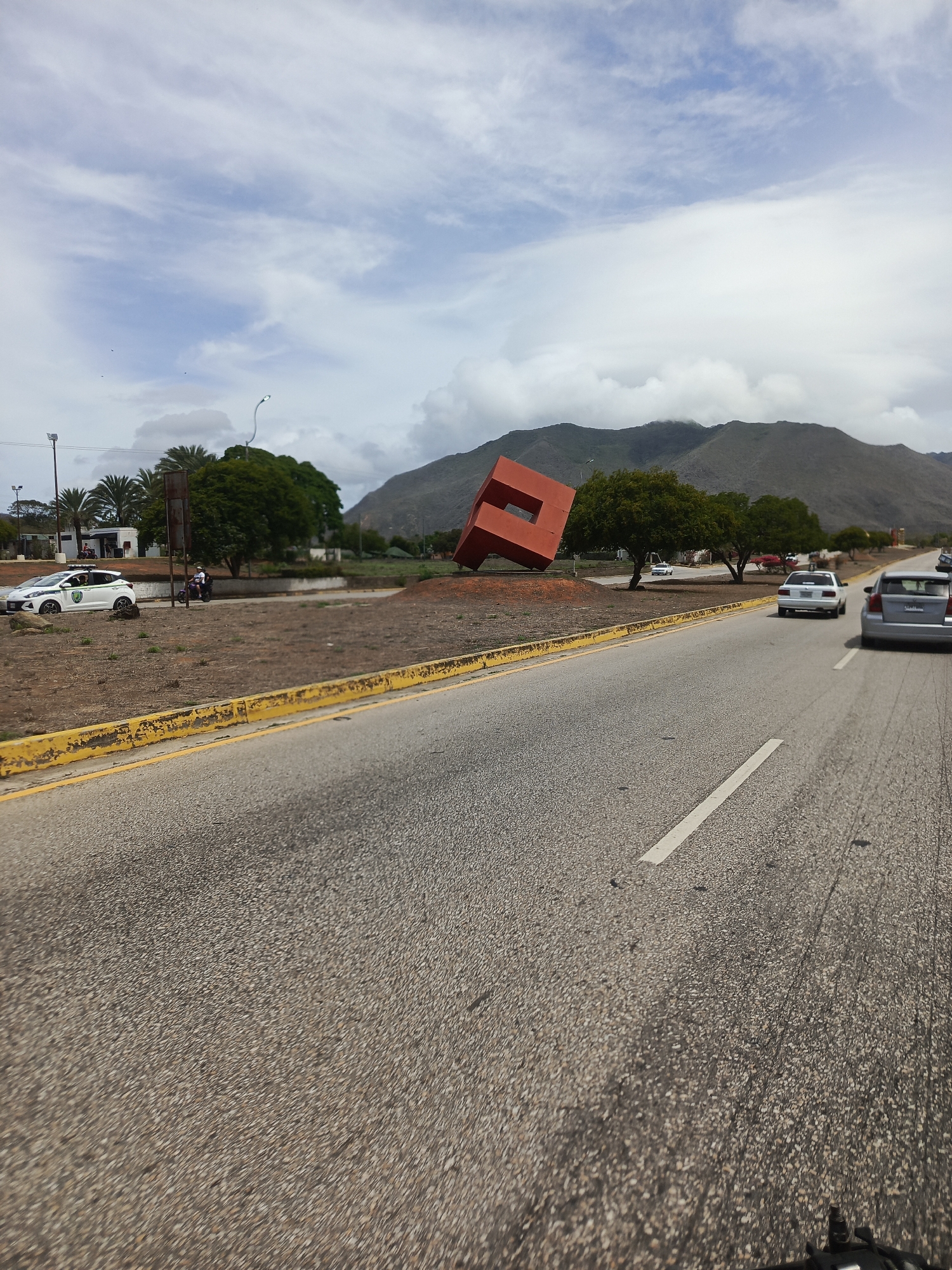 This image depicts a roadside scene in Los Cerritos, Venezuela, under a cloudy sky. A two-lane asphalt road occupies the foreground and mid-ground, featuring a solid yellow curb line and a white dashed lane marker. A dry, unpaved median strip, sparsely covered with brown vegetation, separates the road from the far left area.
A prominent red sculpture, geometrically configured as a tilted cubic form with a large square cut-out, stands on the median strip. Two green leafy trees are positioned behind the sculpture. To the left of the sculpture, a white four-door sedan, identified as a police vehicle, is parked, facing left. A lone individual is discernible near the rear left side of this police vehicle. Closer to the sculpture, a dark-colored motorcycle, carrying two occupants, is stationary on the median strip.
Further down the road, a white sedan and a grey sedan are visible in the right lane, moving away from the viewer. In the far left background, a cluster of palm trees and several small white buildings are present. A range of mountains, partially obscured by clouds and covered with green foliage, dominates the background. The sky above is largely covered with white and light blue clouds.
