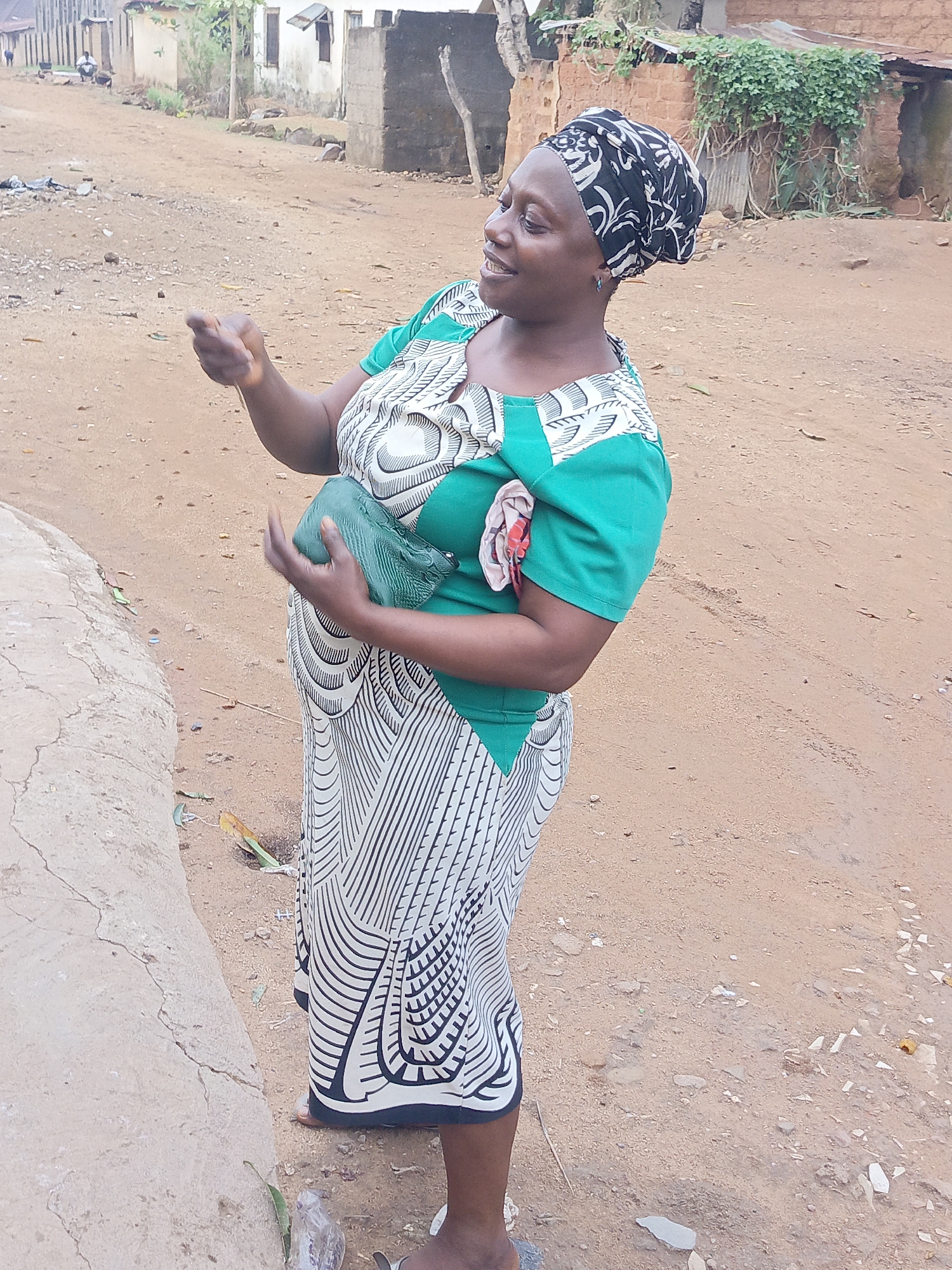 The image captures a woman in Akwanga, Nigeria, standing on a dirt ground. She is wearing a patterned dress with green sleeves and a black and white headscarf. She holds a green object in her hands and is gesturing with her other hand. The background shows modest buildings and a dirt road, typical of a rural setting.