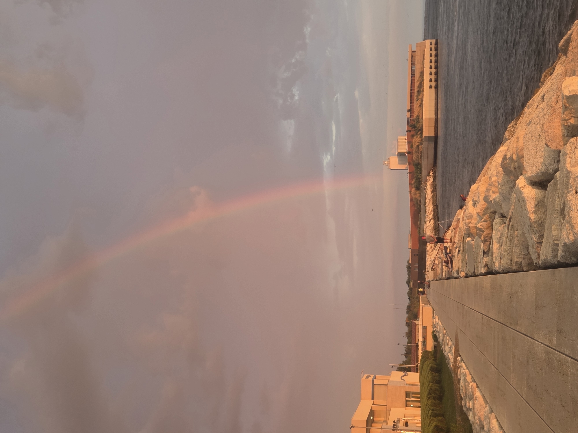 The image depicts a coastal scene in Chioggia, Italy, with a prominent rainbow arching across a muted sky. The foreground features a paved walkway bordering a rocky breakwater. Several individuals are present, some appearing to be fishing from the rocks. On the left side of the walkway, a modern building with cream-colored facades and blue-tinted windows is visible, bordered by manicured greenery. Further in the distance, along the coastline, another long, low-lying building with a contemporary design, featuring a brown roof and large windows, is situated. The sea, a deep blue, gently laps against the rocky shore. The overall lighting suggests either dawn or dusk, casting a warm, reddish-orange hue over the scene, particularly on the rocks and the sky.
