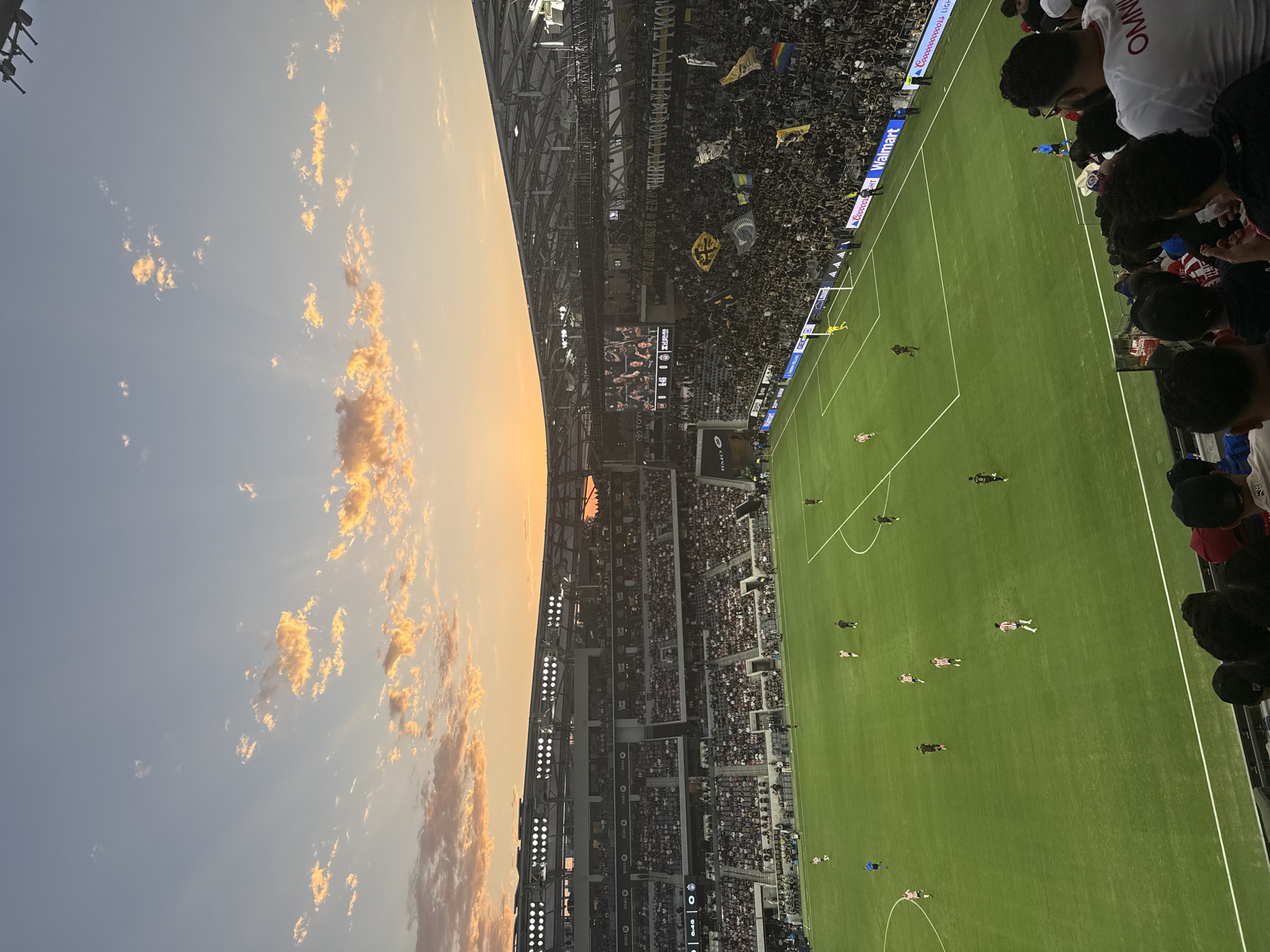 This image captures a sunset over a soccer stadium in Los Angeles, California, United States. The sky is painted with hues of orange and yellow, with scattered clouds catching the golden light. The stadium is filled with spectators, many wearing dark clothing, creating a dense sea of fans. The green soccer field is visible below, with several players in action, and the stadium's bright lights are illuminated. Large screens display game information and images of fans. Banners with text like "THIRTY TWO IN FIFTY TWO" are displayed within the stadium. The overall scene suggests a lively sporting event taking place at dusk.