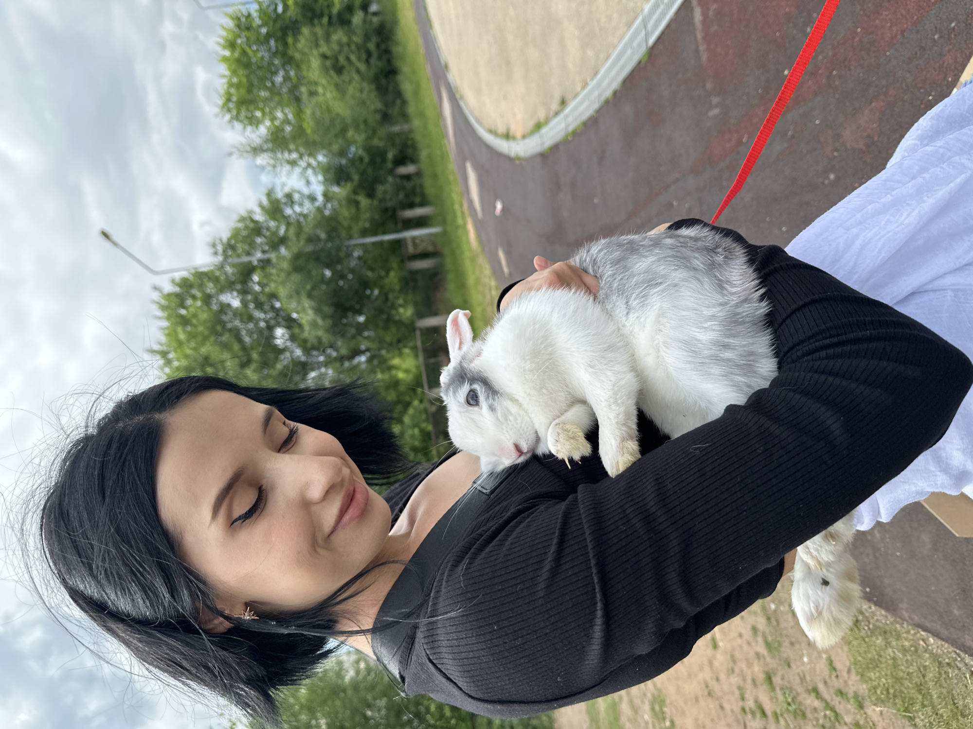 In an outdoor setting, a young woman with black hair and light skin is holding a rabbit. She is wearing a black, ribbed long-sleeved shirt. Her eyes are closed, and she has a gentle smile on her face, conveying a sense of peace and contentment. The rabbit, which is predominantly white with gray markings on its head and back, is nestled in her arms, its head resting on her shoulder. A red leash is attached to the rabbit.
The background suggests a park or recreational area with a paved path curving into the distance. Lush green trees and vegetation are visible, indicating a pleasant day. The sky appears overcast, with patches of white clouds. The overall impression is one of a serene moment shared between the woman and her pet. The time of day is likely daytime, given the ambient light. The location is identified as Chaikovsky, Russia. There is no visible text in the image.