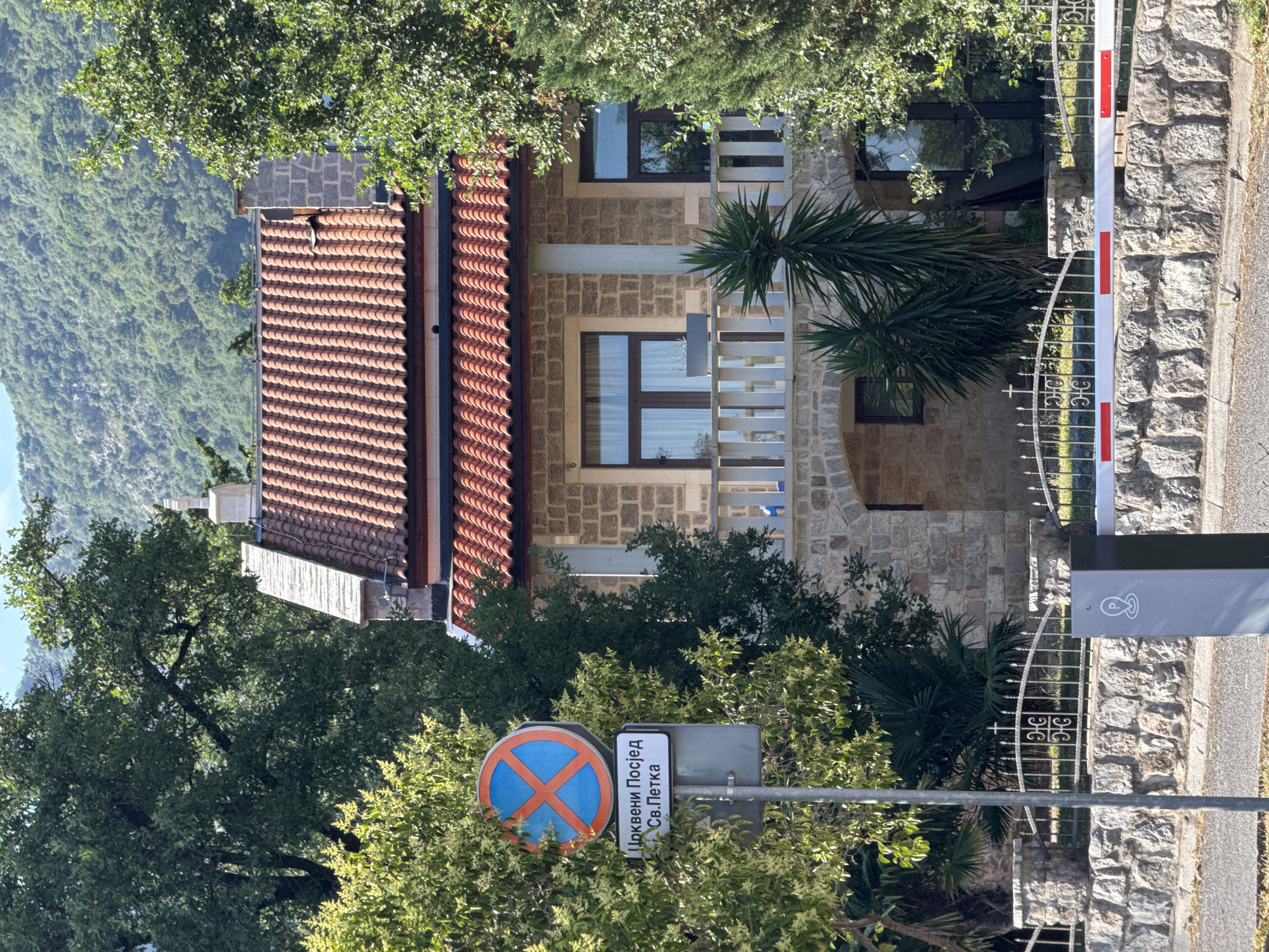 The image depicts a street scene in Budva, Montenegro. The foreground features a street sign indicating "Црквени Посиед Св. Петка", which translates to "Church Visit St. Petka". Next to it is a circular traffic sign with a blue background and a red cross, signifying no stopping. A white barrier with red stripes is also visible, suggesting a controlled entry or exit.
Behind the signs, a building constructed with stone blocks and featuring a red-tiled roof is partially obscured by lush green trees and palm fronds. The building has several windows, some reflecting the sky. A wrought-iron gate with decorative elements stands in front of the building. In the background, a steep, tree-covered mountain slope rises, indicating a hilly or mountainous terrain. The time of day appears to be daytime, with bright sunlight casting shadows, and the weather seems clear. There are no people visible in the image. The scene conveys a sense of tranquility and a picturesque setting typical of a Mediterranean coastal town.