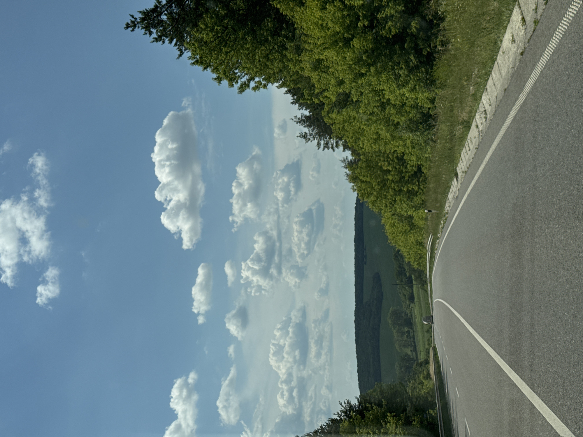 This is a vertical, high-angle shot of a paved road curving to the right, bordered by a grassy verge and a stone retaining wall on the left. Lush green trees form a dense canopy along the left side of the road, with their branches extending into the frame. The sky above is a vibrant blue, dotted with fluffy white cumulus clouds. In the distance, a rolling landscape of green fields and more trees can be seen under the bright sky. The road itself has a white line marking its edge. The lighting suggests it is daytime, likely late morning or early afternoon, with clear, sunny weather. The perspective gives a sense of movement, as if the viewer is driving along the road. There are no visible people or text in the image. The scene evokes a feeling of a pleasant, scenic drive through a rural or semi-rural area.