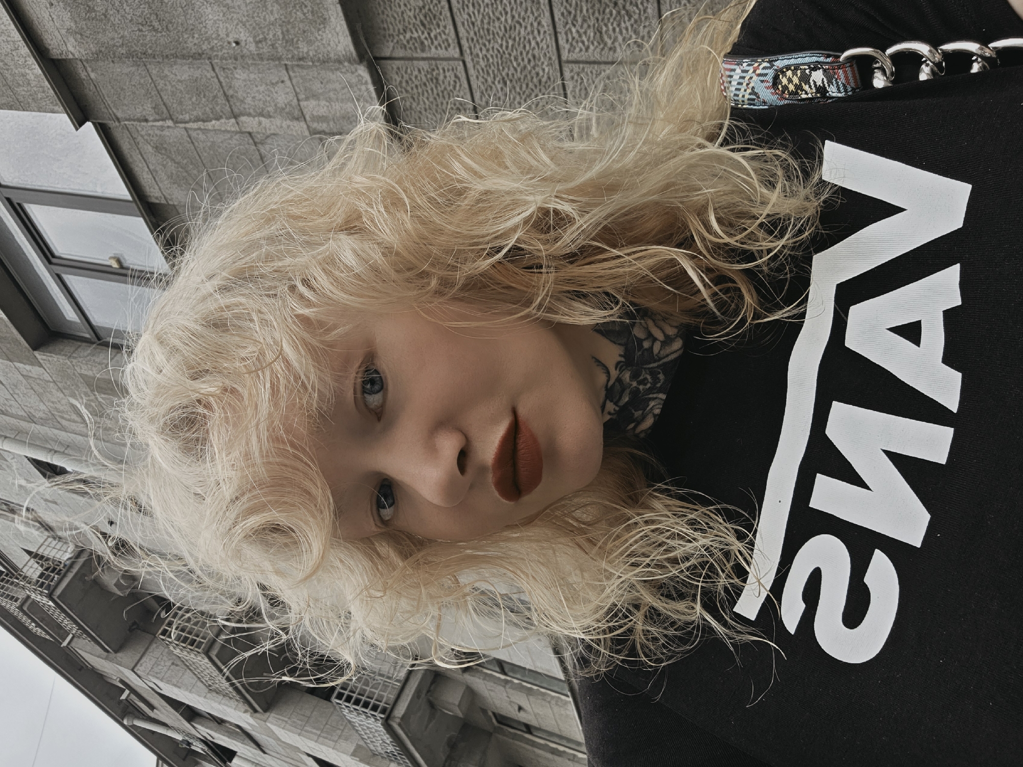A close-up selfie depicts a female subject with light blonde, wavy hair and blue eyes, looking directly at the camera. She wears red lipstick and has a black, intricate tattoo visible on the left side of her neck. Her attire consists of a black t-shirt with a mirrored white "VANS" logo. A colorful, patterned strap with silver chain links, suggesting a bag, is positioned over her right shoulder. The background features a tall, multi-story building, characteristic of architecture found in Saint Petersburg, Russia. The building's facade is gray, constructed from stone or concrete, and includes multiple balconies with metal railings and dark-framed windows. The sky is overcast and gray.