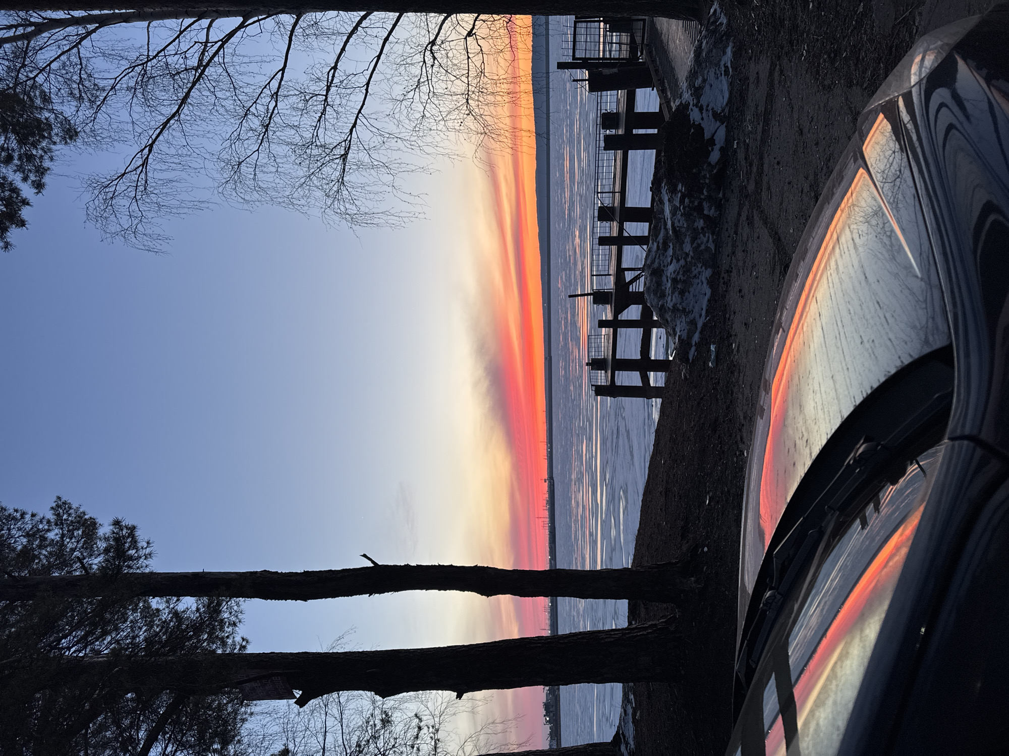 The image shows a sunset scene in what appears to be the city of Chaikovsky, Russia. The sky is a gradient of colors, ranging from blue to orange and red. A frozen body of water stretches into the distance, reflecting the colors of the sunset. A wooden pier extends into the water. In the foreground, the edge of a car is visible, with the sunset reflected in its surface. Trees frame the scene.