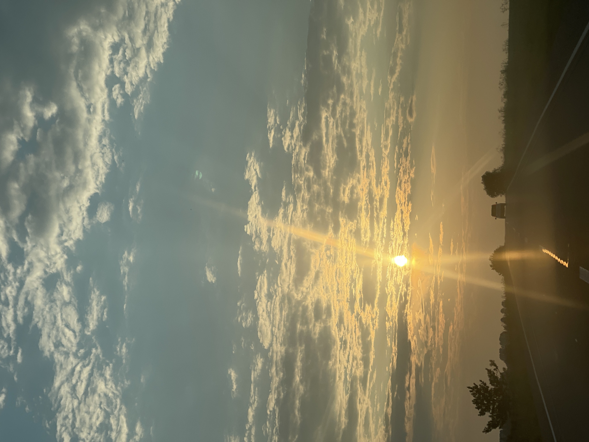 The image shows a road with two cars driving away from the viewer. The sun is setting, casting a golden light over the scene. The sky is filled with clouds, and the landscape is dotted with trees and fields. This scene is located in the city of Perevoznoye, Russia.