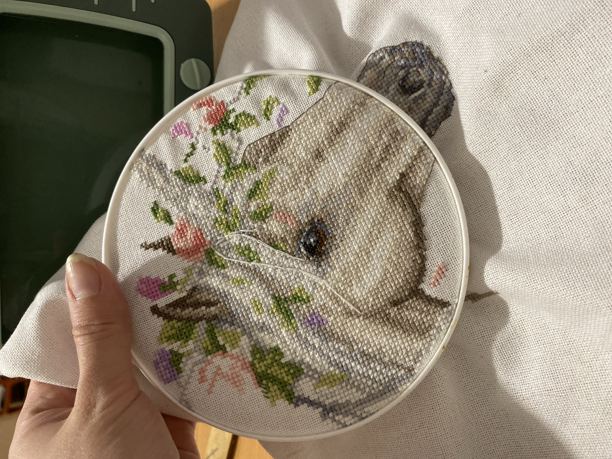 A person holds an embroidery hoop containing a cross-stitched image. The design features a stylized animal, likely a rabbit or hare, surrounded by floral elements. The cross-stitch is in progress, with a needle and thread visible in the eye area. The embroidery is mounted on a larger piece of white fabric. Part of a green object, possibly a lamp or other device, is visible in the upper left corner. No information about the location in Yekaterinburg, Russia, can be discerned from the image.