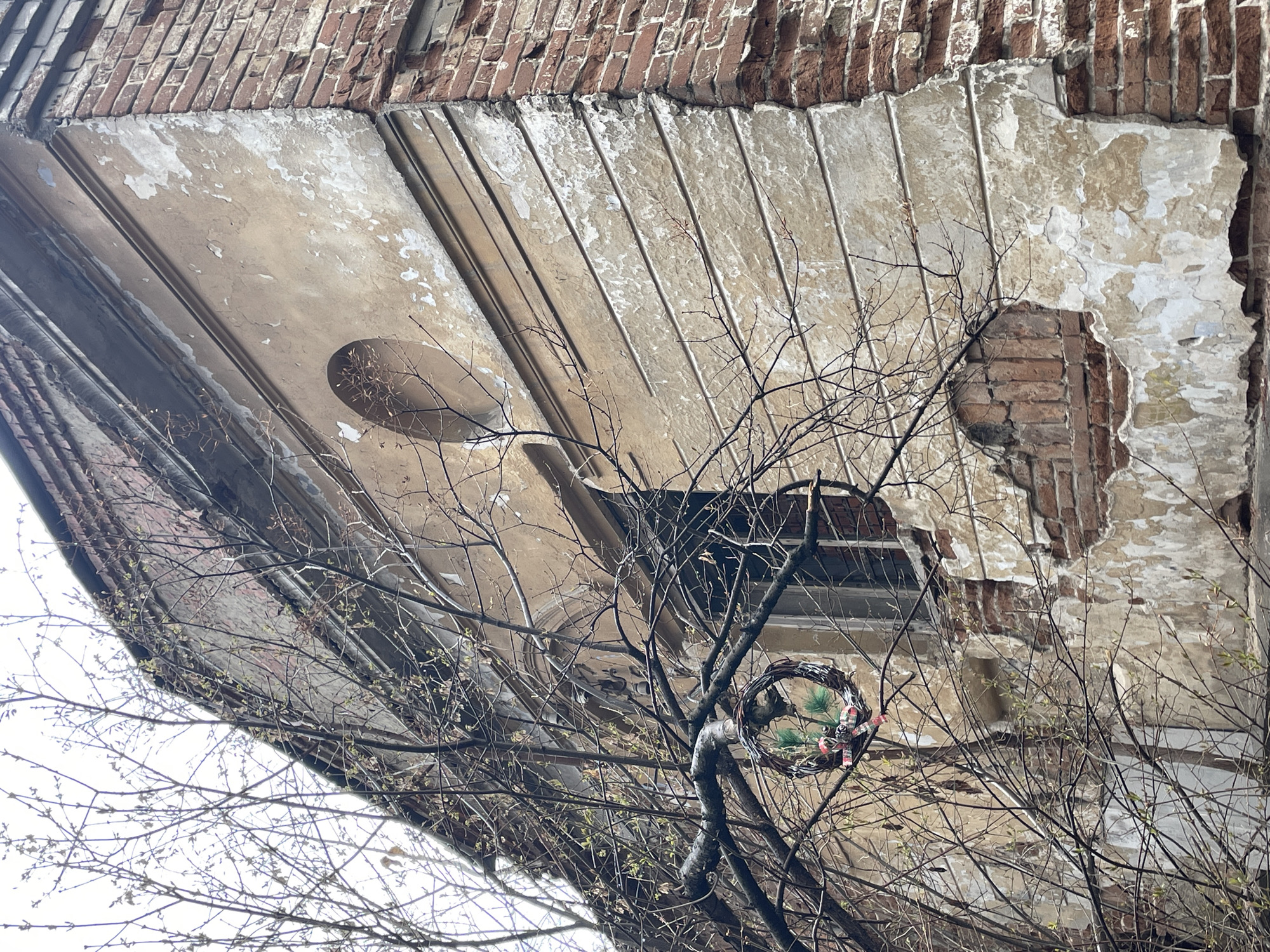 This low-angle shot captures the underside of a severely dilapidated building structure, likely a balcony or cornice, framed by a network of bare tree branches. The setting suggests an urban environment, specifically Yekaterinburg, Russia, as provided by the context.
The building element features a base of reddish-brown brickwork that is exposed in several areas due to extensive decay. Beneath this, a layered facade or soffit, initially light beige or off-white, shows significant damage. Large sections of plaster or paint have peeled away, revealing a rough, textured surface, possibly concrete, with decorative vertical or diagonal grooves. A prominent, irregular hole near the center-right exposes a distinct pattern of reddish-brown bricks, indicating the underlying construction. Other patches of exposed brick and crumbling material are visible across the structure.
In the foreground, a dense tangle of dormant tree branches dominates the lower half and parts of the mid-ground. Many branches are bare, characteristic of late winter or early spring, though some tiny green buds are discernible, hinting at nascent growth. Hanging from a thicker branch to the right of center is a small, handcrafted wreath. It appears to be made of natural twigs, adorned with a few green faux pine needles and a small bow tied with red and white striped ribbon, providing a touch of domesticity amidst the decay.
The lighting is soft and diffused, suggesting an overcast sky, with no harsh shadows. No human activity is depicted. The scene evokes a sense of neglect and the passage of time, juxtaposed with the subtle signs of nature's renewal. There is no visible text in the image.