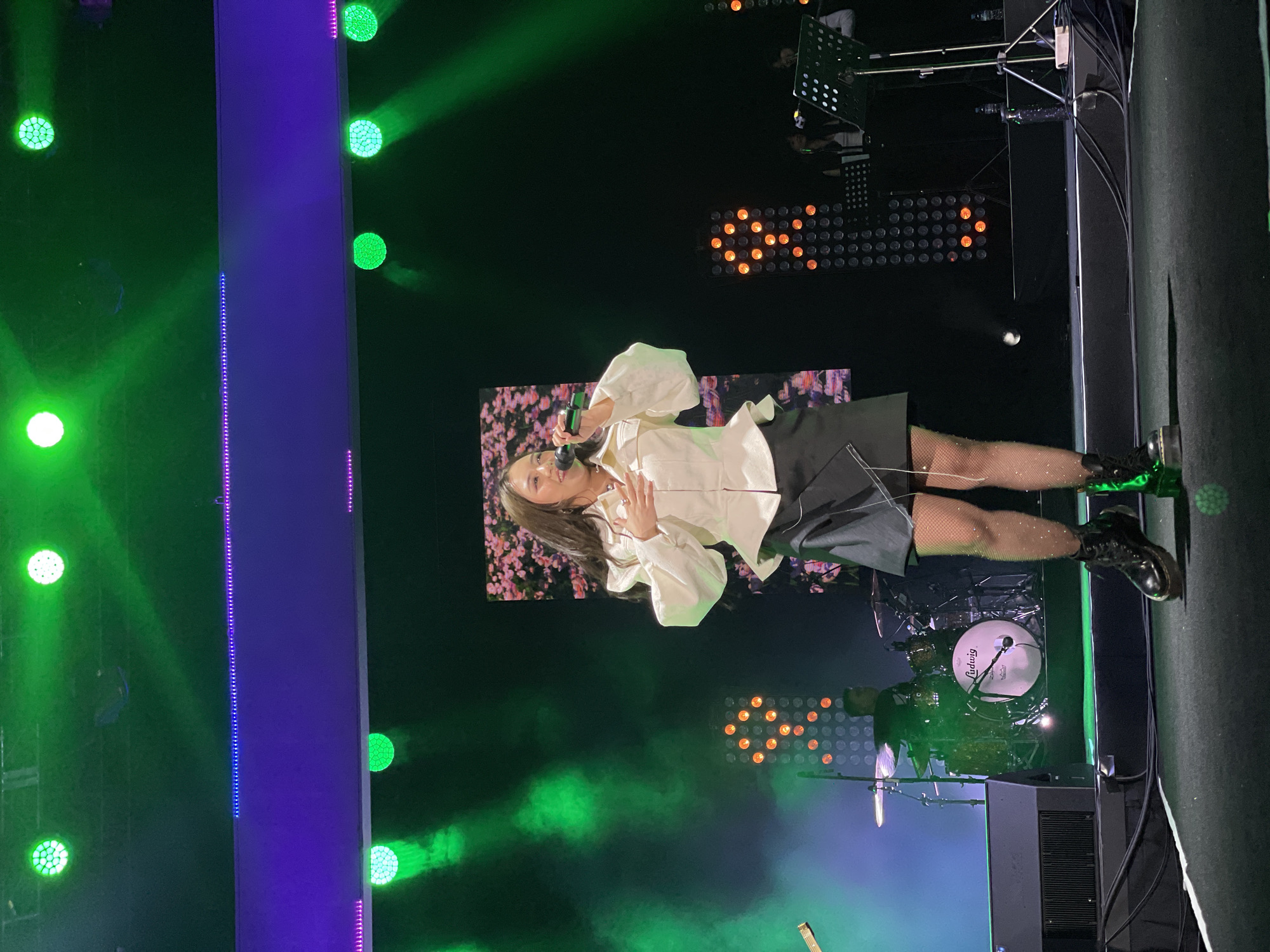 A woman with brown hair is performing on a stage. She is wearing a white blouse with puffed sleeves and a black skirt. She is holding a microphone and singing. Behind her is a screen with a floral pattern. There are green lights and orange lights on the stage. There is a drum set to her right. The location is Salembaran, Indonesia.