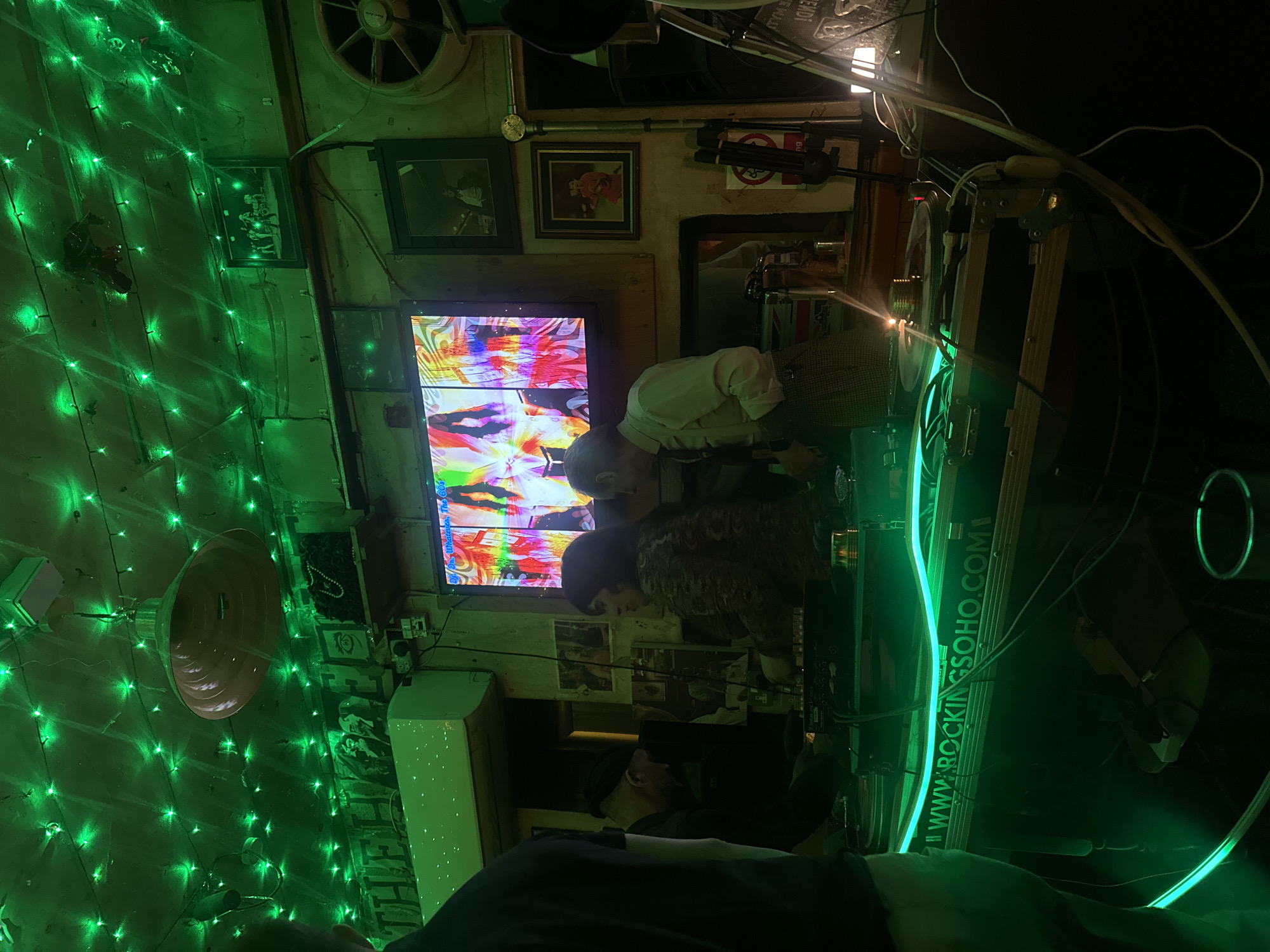 Indoor view of what appears to be a music venue or pub, decorated with green fairy lights and various framed images and objects on the walls. A television screen displaying abstract colorful patterns is prominent. Two individuals are standing close to the screen, possibly watching. The location appears to be in London, United Kingdom, based on the URL "WWW.ROCKINGSOHO.COM" that is visible in the image.