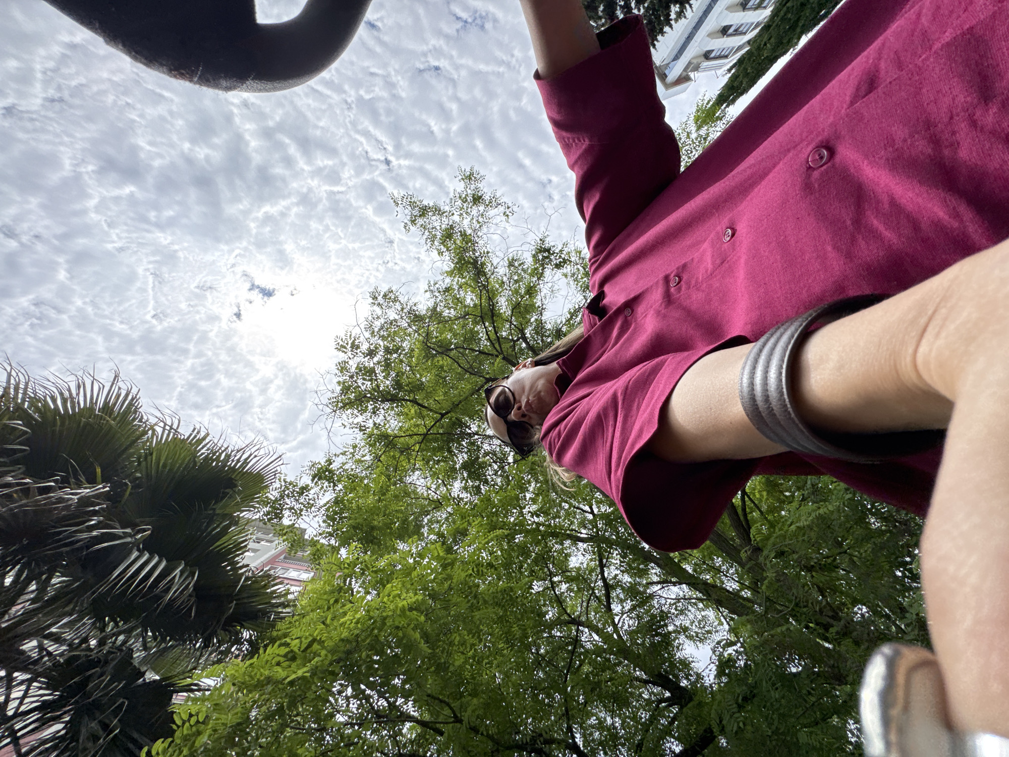 The image is a low-angle, full-body shot of a person holding a camera and taking a selfie in an outdoor setting. The person is wearing a magenta button-down shirt and sunglasses. A thick, dark, possibly metal object, is partially visible at the top of the frame, obscuring some of the sky. The sky is overcast with a textured, cloudy pattern. Lush green trees and palm fronds fill the lower portion of the frame, with a building visible in the background through the foliage. The lighting suggests it is daytime. The overall impression is of a casual, outdoor moment.