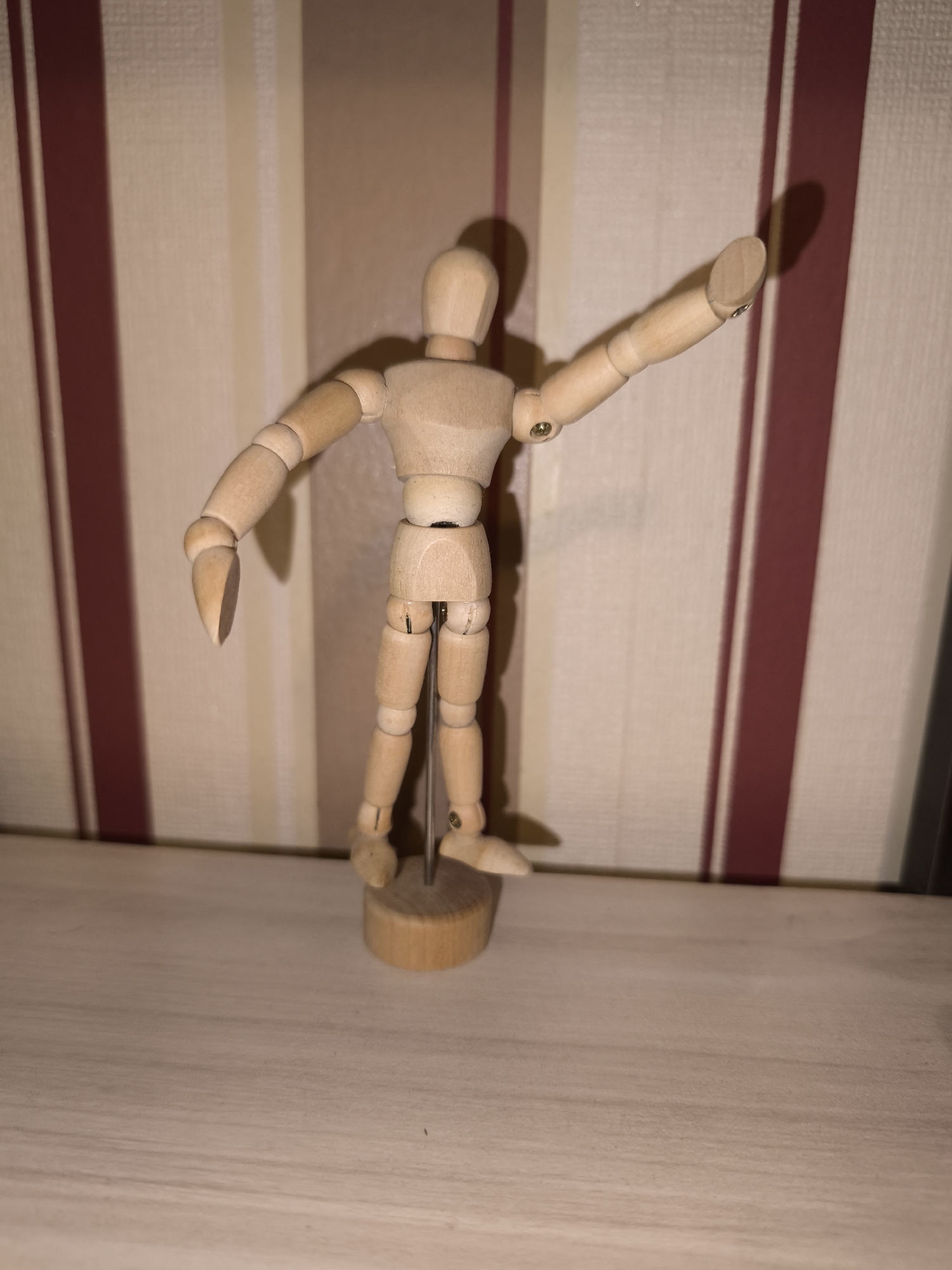 A wooden artist's mannequin stands on a light-colored surface, likely a shelf or table. The mannequin is in a pose with one arm raised upwards and to the right, and the other arm extended slightly downwards and to the left. The mannequin is standing upright and is mounted on a circular wooden base with a metal rod. The background features a vertically striped wall, with alternating light and dark brown stripes. The scene is illuminated by a light source from an unspecified location, casting shadows behind the mannequin. No people are visible. The location in Burgas, Bulgaria is not discernible from the visual information provided.