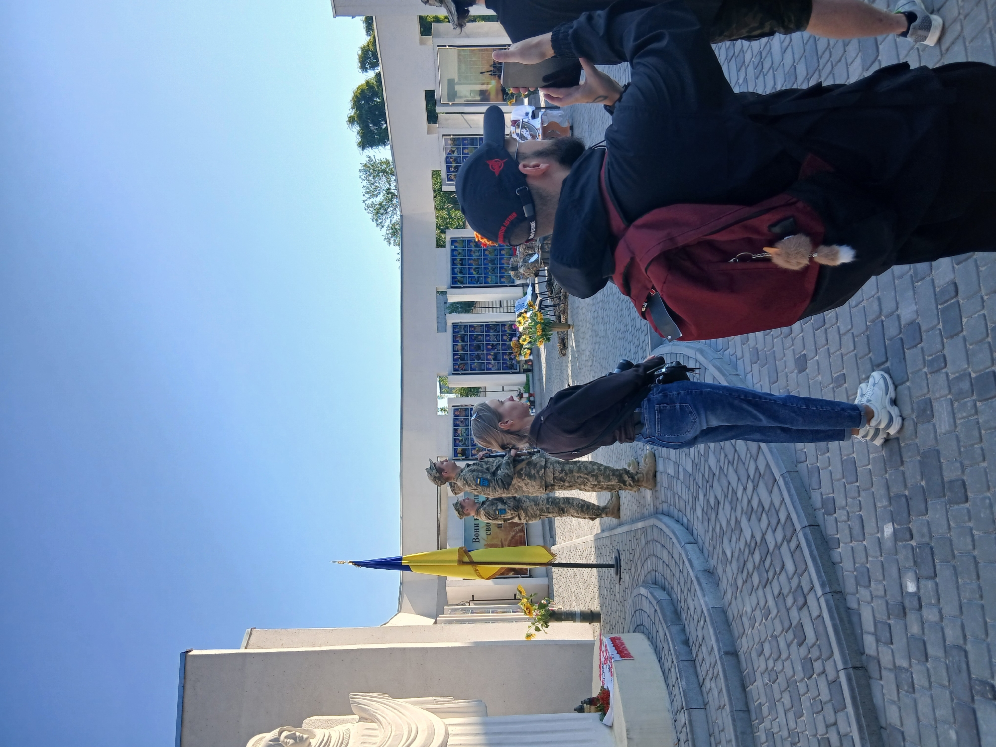 This is a medium shot, taken outdoors, of a memorial or monument site in Verkhnodniprovsk, Ukraine, under a clear blue sky. In the foreground, a man wearing a black hoodie and a dark baseball cap with red lettering is holding up a smartphone to take a picture. He has a red backpack on his shoulders with a fluffy keychain attached. To his left, a woman in a black top and blue jeans stands with her back partially turned, looking towards the center of the scene.
In the mid-ground, several individuals in Ukrainian military uniforms are standing at attention. One soldier is holding a rifle. A Ukrainian flag, yellow and blue, is planted near them. Behind the soldiers, a white building with several rectangular panels displaying what appear to be portraits is visible. Sunflowers are arranged in vases in front of the building. The ground is paved with cobblestones, and there are stone steps leading up to a curved platform. A statue or monument with a draped figure is visible on the left side of the frame. The overall atmosphere suggests a solemn commemoration or ceremony.