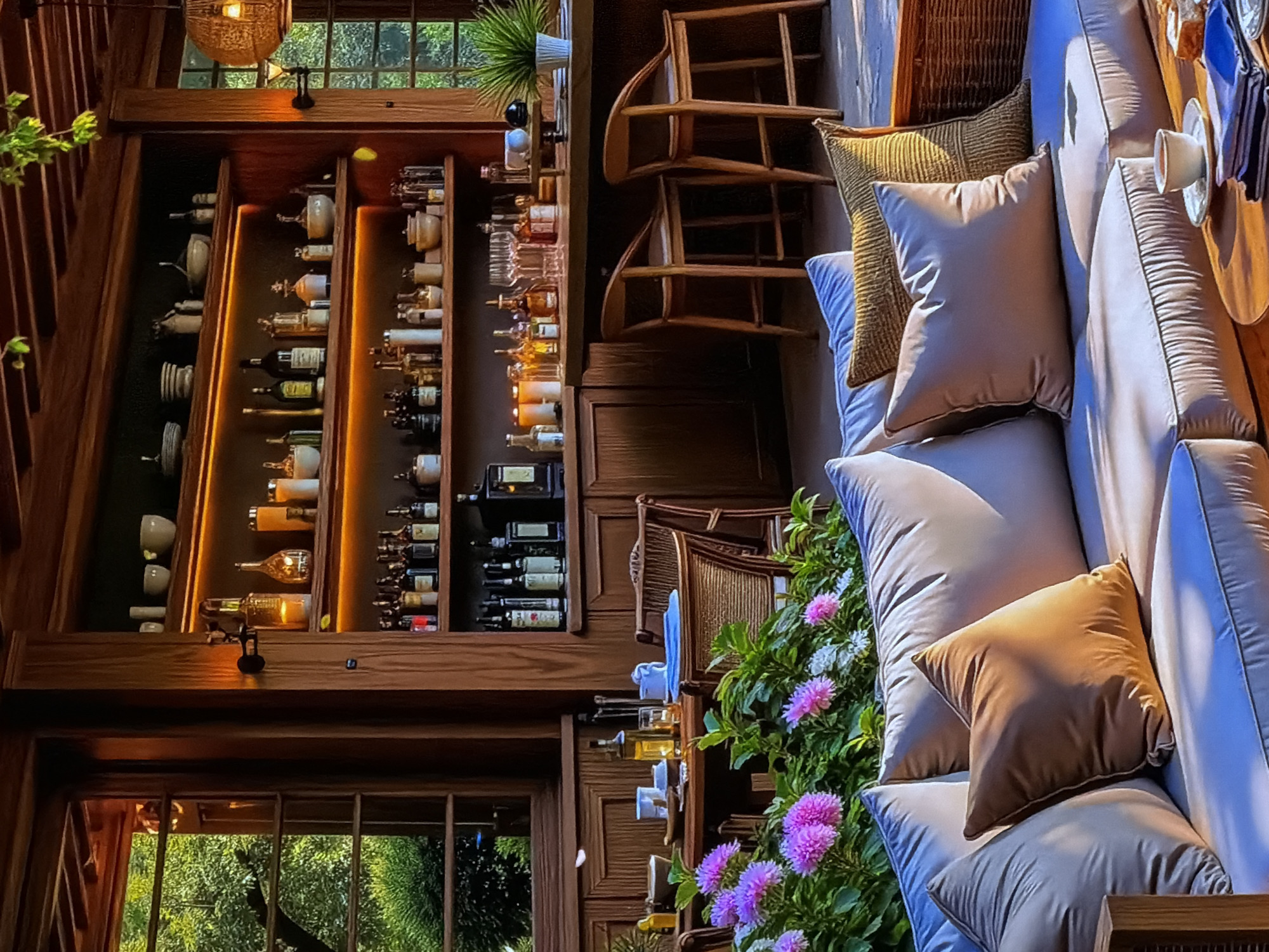 This image captures a warm and inviting indoor setting, likely a lounge area, bar, or restaurant, possibly located in Sofiivska Borshchahivka, Ukraine. The scene is characterized by rich wooden interiors, comfortable seating, and tasteful decor, evoking a sense of cozy sophistication.
On the left side of the frame, a tall, multi-tiered wooden shelving unit serves as a prominent feature. It is illuminated by warm, golden strip lighting that highlights numerous bottles, likely containing spirits or wine, alongside various elegant ceramic bowls and decorative pieces. Above the shelving, a rustic hanging light fixture adds to the ambiance, and through a window, lush green foliage is visible, suggesting a natural outdoor environment.
The right side of the image reveals a comfortable seating arrangement. A long, light-colored sofa or bench is adorned with several plush cushions, including soft beige and a textured golden pillow. Adjacent to the sofa, a wooden surface holds a white teacup and saucer, a folded napkin, and other small items, suggesting a moment of quiet relaxation or anticipation of guests. Behind the sofa, two wooden chairs with woven backs are visible, further contributing to the natural, rustic-chic aesthetic. In the foreground on the right, a vibrant cluster of green plants featuring purple and white dahlia-like flowers adds a refreshing burst of color to the indoor space.
No people are present in the image, but the arrangement of objects suggests a space designed for leisure and social interaction. The interplay of warm artificial lighting and the natural light filtering through the windows creates a soft, inviting atmosphere, hinting at either late afternoon or early evening. No text is visible in the frame.