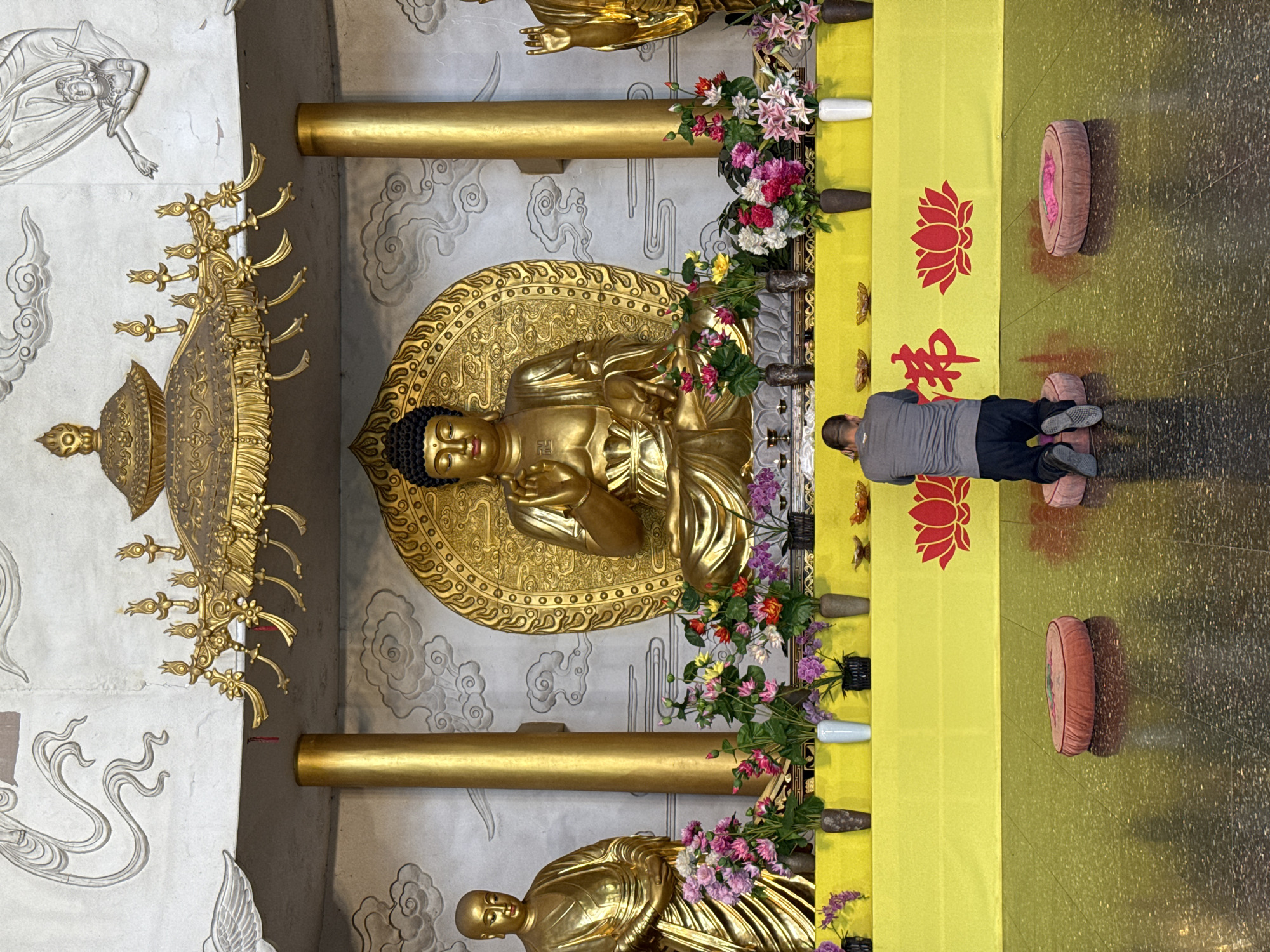 This image captures the interior of a Buddhist temple or shrine in Shizhong, China. The central focus is a large, golden Buddha statue, seated in a meditative posture on an intricately detailed golden pedestal, backed by a circular, halo-like structure. The Buddha has a serene expression, dark hair, and a small red mark on its forehead. Partially visible on either side are two other golden statues of Buddhist figures. Above the left-most statue hangs an ornate golden canopy.
In the foreground, a man dressed in a grey t-shirt and dark pants kneels on a pink cushion, facing the central Buddha statue in an act of prayer or reverence. Another pink kneeling cushion rests on the polished, dark floor nearby. A long yellow fabric runner stretches across the floor in front of the statues, adorned with red lotus flower symbols and two red Chinese characters that read "佛殿" (Fódiàn), signifying "Buddha Hall."
The temple walls are light-colored and decorated with subtle embossed patterns depicting clouds and celestial figures. Golden pillars accent the space. Numerous vases filled with artificial flowers in shades of pink, purple, red, and yellow are arranged along the yellow runner, adding color to the predominantly golden and muted palette. The scene conveys a tranquil and devotional atmosphere.