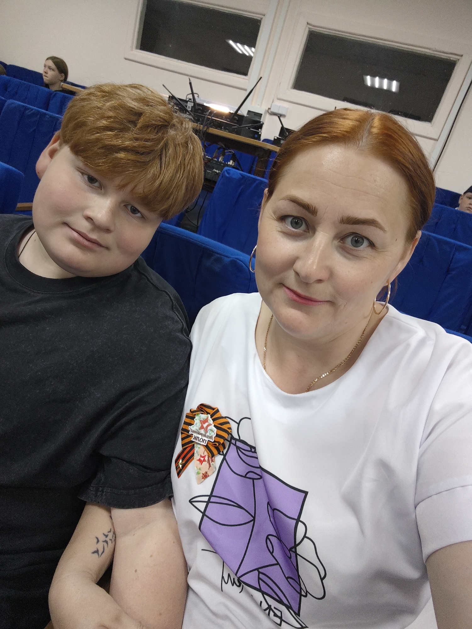A photograph depicts a red-haired woman and a red-haired boy in an indoor setting. The woman is positioned on the right, wearing a white t-shirt with a stylized purple design. She has gold hoop earrings and a gold necklace. The boy, on the left, wears a dark t-shirt. Both are seated on blue upholstered chairs.
A St. George ribbon is pinned to the woman's shirt. A tattoo of birds is visible on the woman’s arm.
In the background, there are additional rows of blue seating, and windows. Equipment, possibly audio-visual, is visible behind the first row of seats. The photo appears to have been taken in a theater or auditorium setting.
The location is Фоки, Russia.