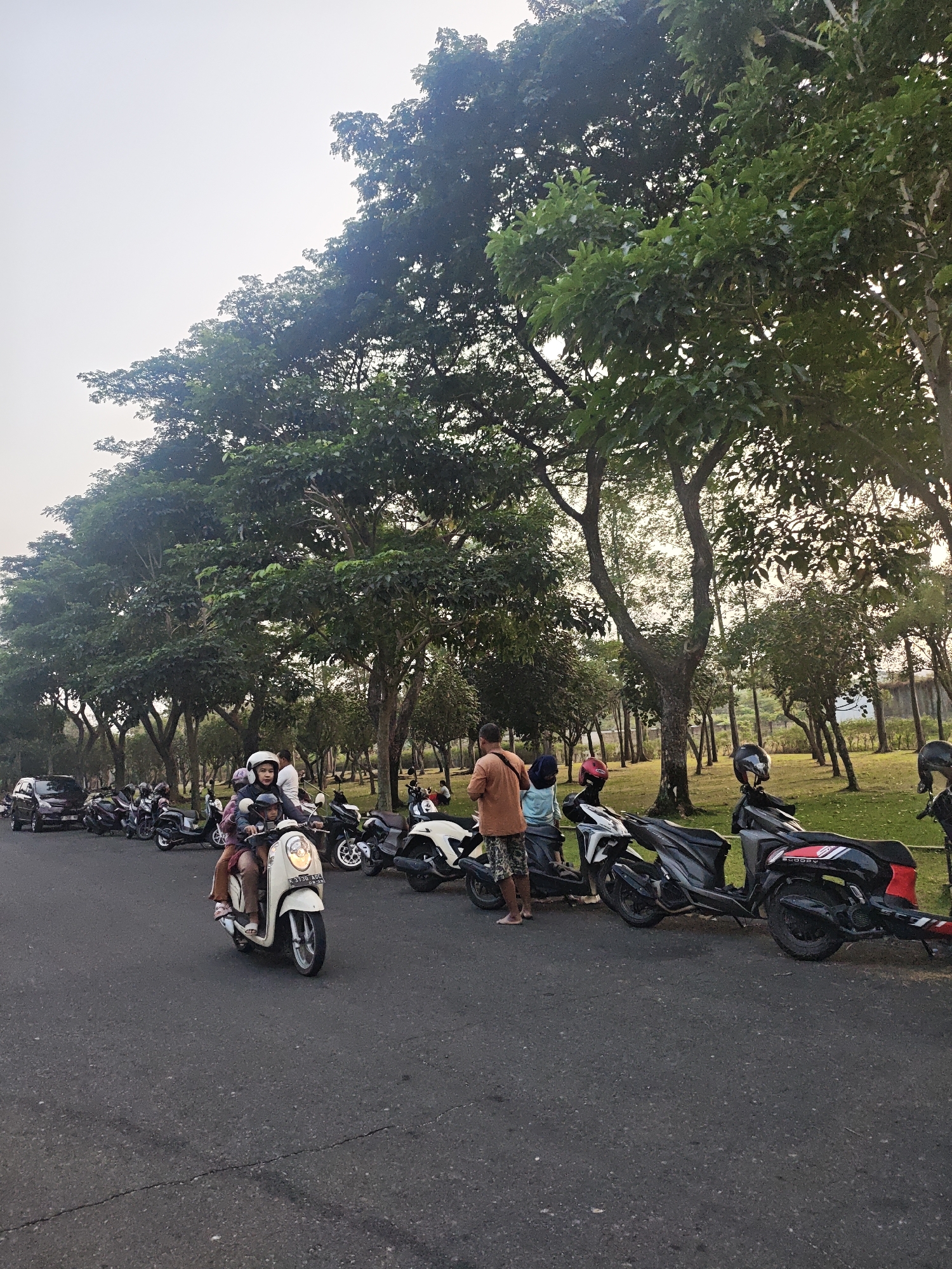 The image depicts an outdoor scene, possibly a park or roadside, with a prominent line of trees on the right side. Several parked motorcycles are visible along the side of an asphalt road. In the foreground, two individuals, appearing to be a young person and an adult, are riding a light-colored scooter. The scooter has a visible license plate. Further down the road, other motorcycles are parked in a row, and a dark-colored car is also present. Several people are standing near the parked motorcycles. The environment appears to be urban or semi-urban, with a hazy sky in the background. The lighting suggests it is either early morning or late afternoon. The location is identified as Bacin, Indonesia.