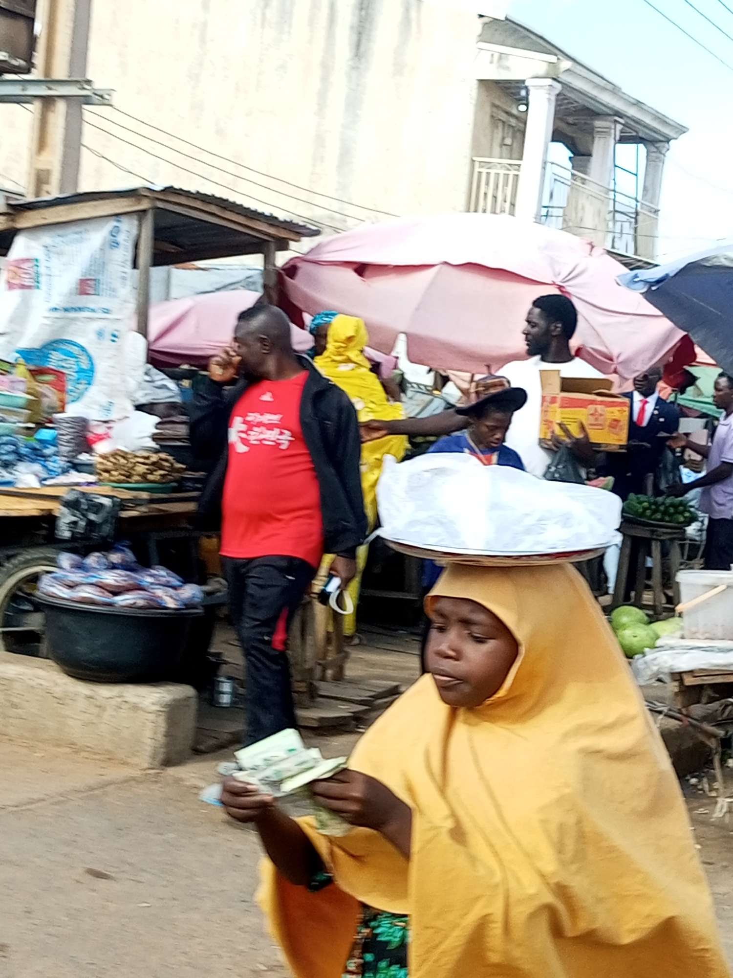 This is a street-level, eye-level, medium shot photograph taken in the city of Tempure, Nigeria, showing a bustling market scene. In the foreground, a young girl wearing a yellow hijab and a patterned skirt balances a large white object on her head, with her hands holding a fan of currency. Her mouth is slightly open, and she appears to be speaking or reacting to something. Behind her, a man in a red shirt and black jacket is walking and gesturing with his right hand near his face. Other individuals are visible in the background, including a person in a yellow hooded garment, another man in a white shirt holding a box, and a man in a suit and tie. The environment appears to be an outdoor market with stalls covered by umbrellas and tents, displaying goods such as bags of items, produce like cucumbers, and what looks like gourds. The ground is dusty, and the overall atmosphere suggests active commerce and daily life.