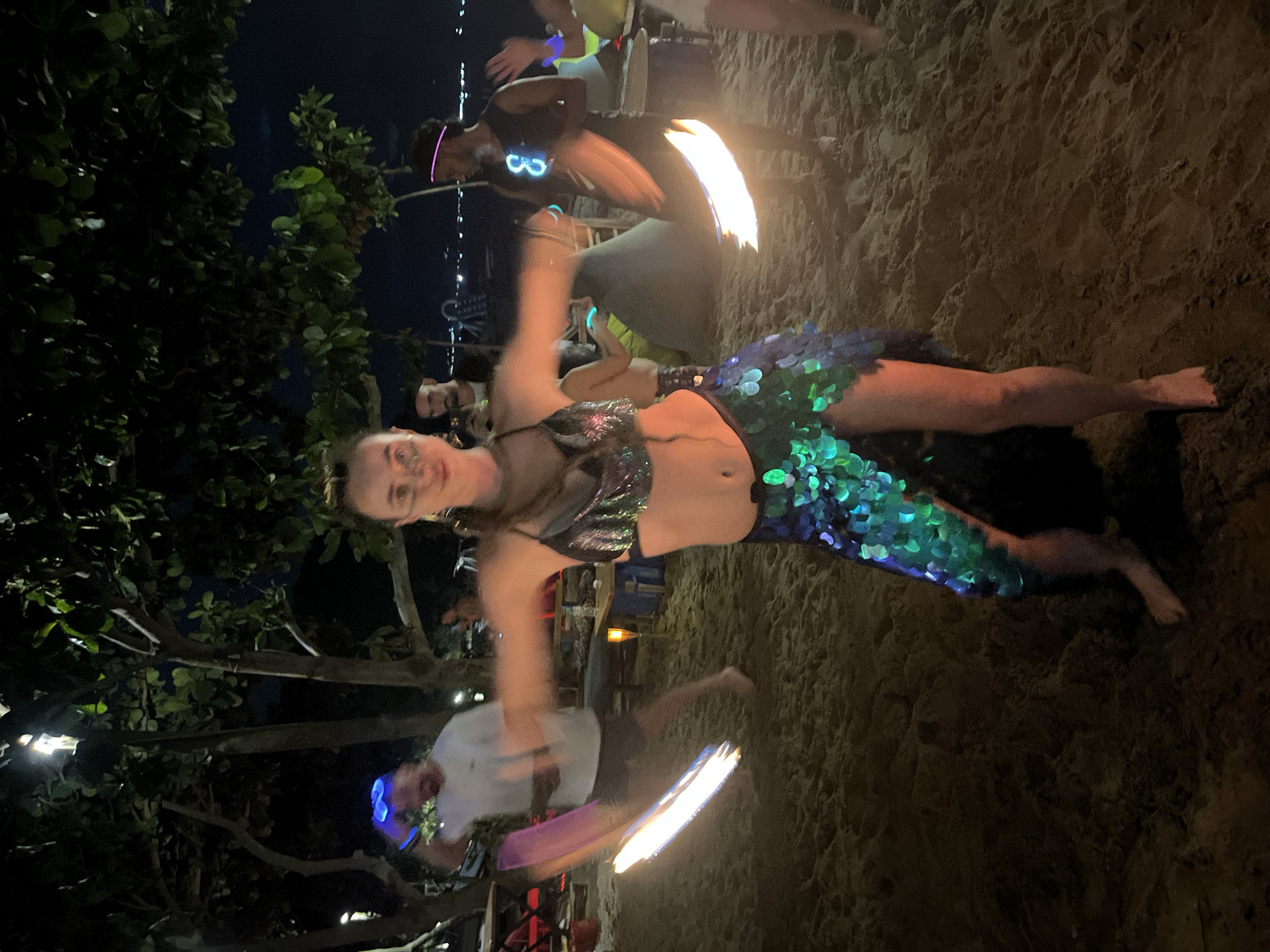 The image depicts a nighttime scene on a sandy beach, likely in Da Nang, Vietnam, with several individuals engaged in fire dancing. The main subject is a woman in the foreground, dressed in a sparkling, sequined outfit that resembles a mermaid's costume, with a bikini top and a scaled skirt. She is captured in motion, her arms blurred by the movement of fire poi, which are creating streaks of light.
Behind her, other performers are also visible, some with glowing accessories like neon headbands and leis. One person in the mid-ground is holding a flaming prop, casting an orange glow on the sand and surrounding foliage. The beach appears to be part of a lively evening gathering, with hints of a bar or seating area with lights in the background. The overall atmosphere is festive and energetic, with the darkness of night contrasted by the bright flames and neon lights. The sand shows footprints and texture, indicating it is a natural beach environment. The presence of trees on the left side of the frame adds to the natural setting. The time of day is clearly night, with the sky being dark and only illuminated by artificial lights and the fire.