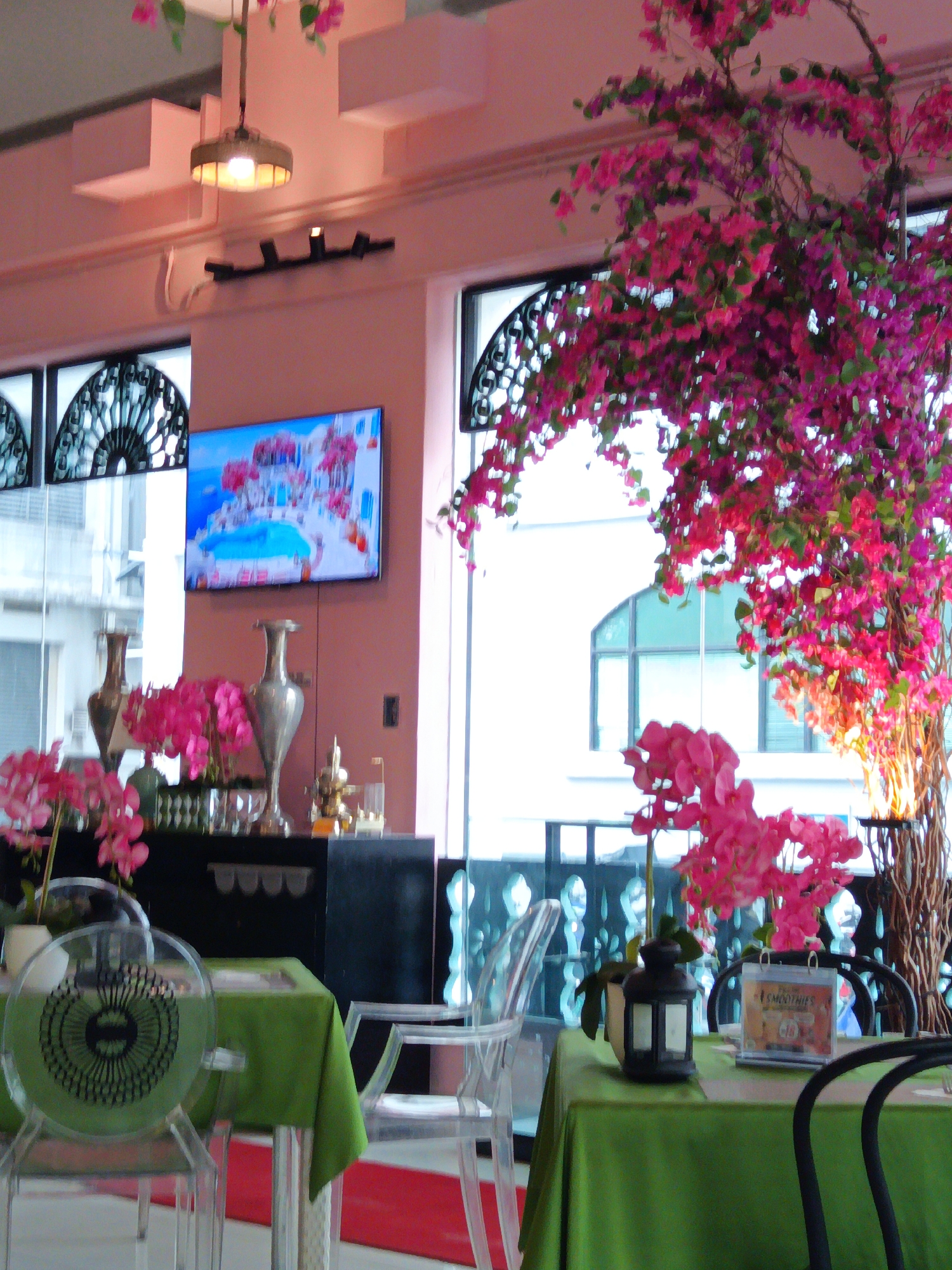 This image depicts the interior of a restaurant in Kuala Lumpur, Malaysia. The scene is set with pink walls and ceiling accents, and large windows offering a view of an urban exterior. Dominating the foreground are dining tables set with green tablecloths and surrounded by clear, modern chairs. Decorative elements include a large artificial tree with vibrant pink flowers, a television screen displaying a scenic image, and ornate vases. The overall atmosphere appears to be chic and airy, possibly indicating a cafe or a restaurant focusing on a bright, floral theme.