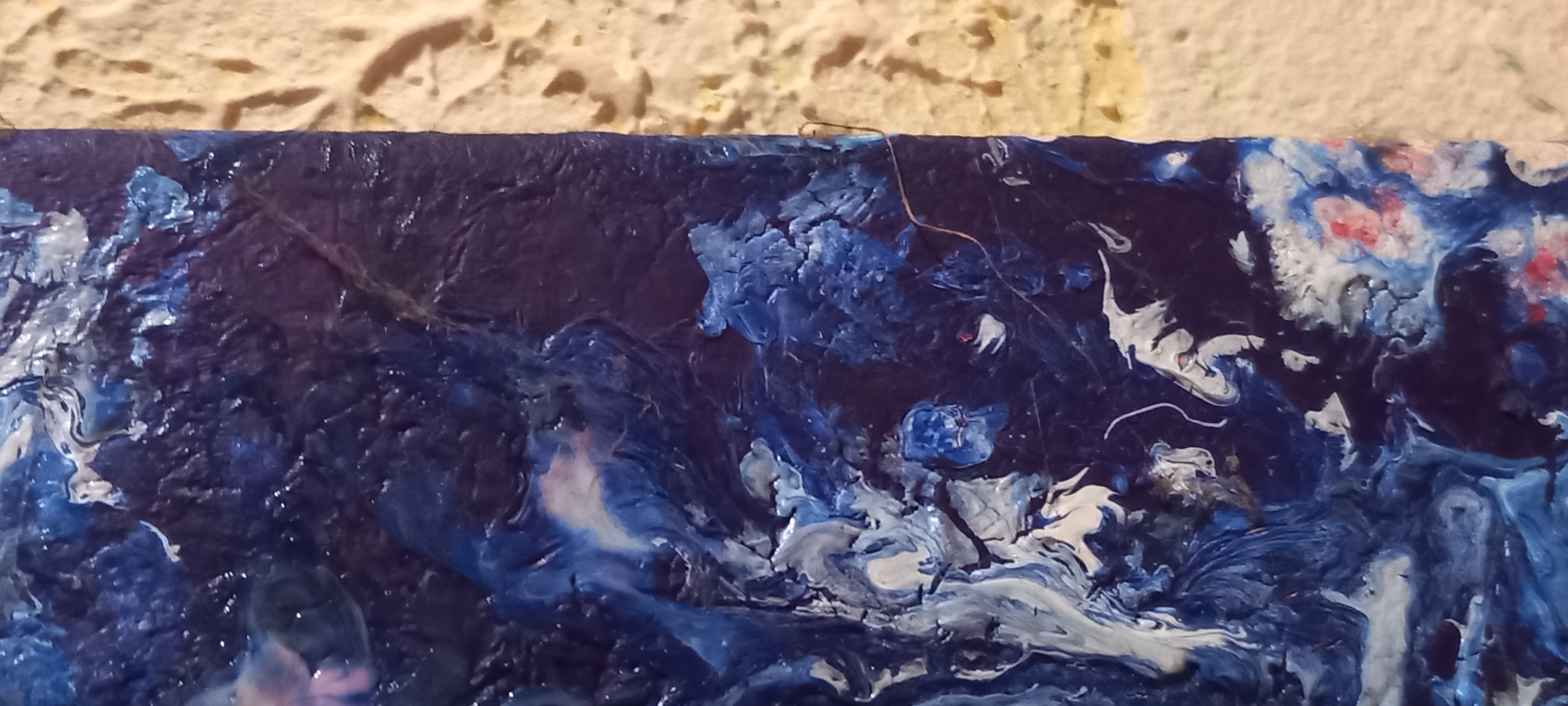 The image displays a highly textured, abstract artwork occupying the left side of the frame, positioned vertically. The artwork features prominent impasto-like swirls and patterns predominantly in dark blue, royal blue, and white, with smaller localized areas showing hints of pink and light red. The surface exhibits a varied texture, ranging from glossy, smooth-looking areas reflecting light to rugged, craggy sections with deep crevices and raised peaks of paint.
To the right of the artwork, a roughly textured, yellowish-beige surface, likely a wall, fills the remainder of the frame. The texture of this surface appears coarse and irregular. A thin, light brown strand, resembling a hair or fiber, stretches horizontally across the dark blue area of the artwork, near the center-right, extending slightly onto the beige wall.
There are no people, explicit subjects, or direct actions depicted. The interaction is limited to the abstract artwork resting against or next to the textured wall. The environment appears to be an indoor setting. The image provides no identifiable geographical markers or contextual information to specify a location in the city of Kryvyi Rih, Ukraine, or any other particular city.