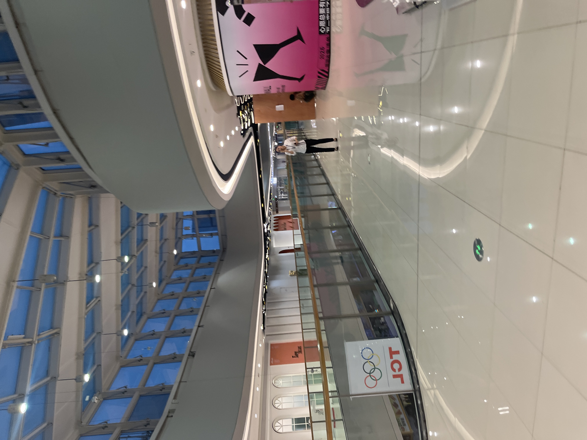 A wide shot captures the interior of a multi-level shopping mall in Shapingba District, China, with a focus on the polished, light-colored tiled floor that reflects the overhead lighting and architectural features. On the left side of the frame, a railing separates the upper level from the lower, where a large sign displaying the Olympic rings and the TCL logo is visible. Further down the upper level, a woman in a white shirt and black pants is walking and holding a phone to her ear. Adjacent to her, a large, cylindrical advertisement with a pink and black graphic of two champagne glasses stands prominently. A second individual, appearing to be a child, is seated near the base of the advertisement. The mall's architecture includes a modern, curved design with a glass ceiling that allows natural light to filter in, illuminating the space and highlighting the clean, contemporary aesthetic.