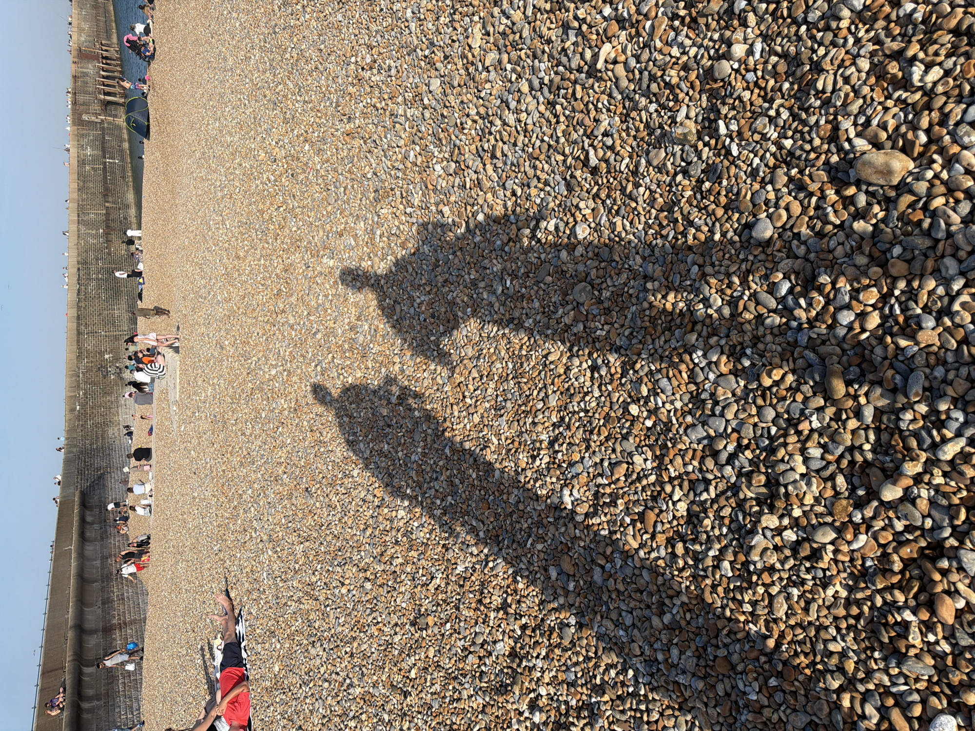 The image shows a pebble beach with people sunbathing and walking along the shoreline, located in Folkestone and Hythe District, United Kingdom. A sea wall runs along the left side of the frame. The shadows of two individuals are cast prominently in the foreground.