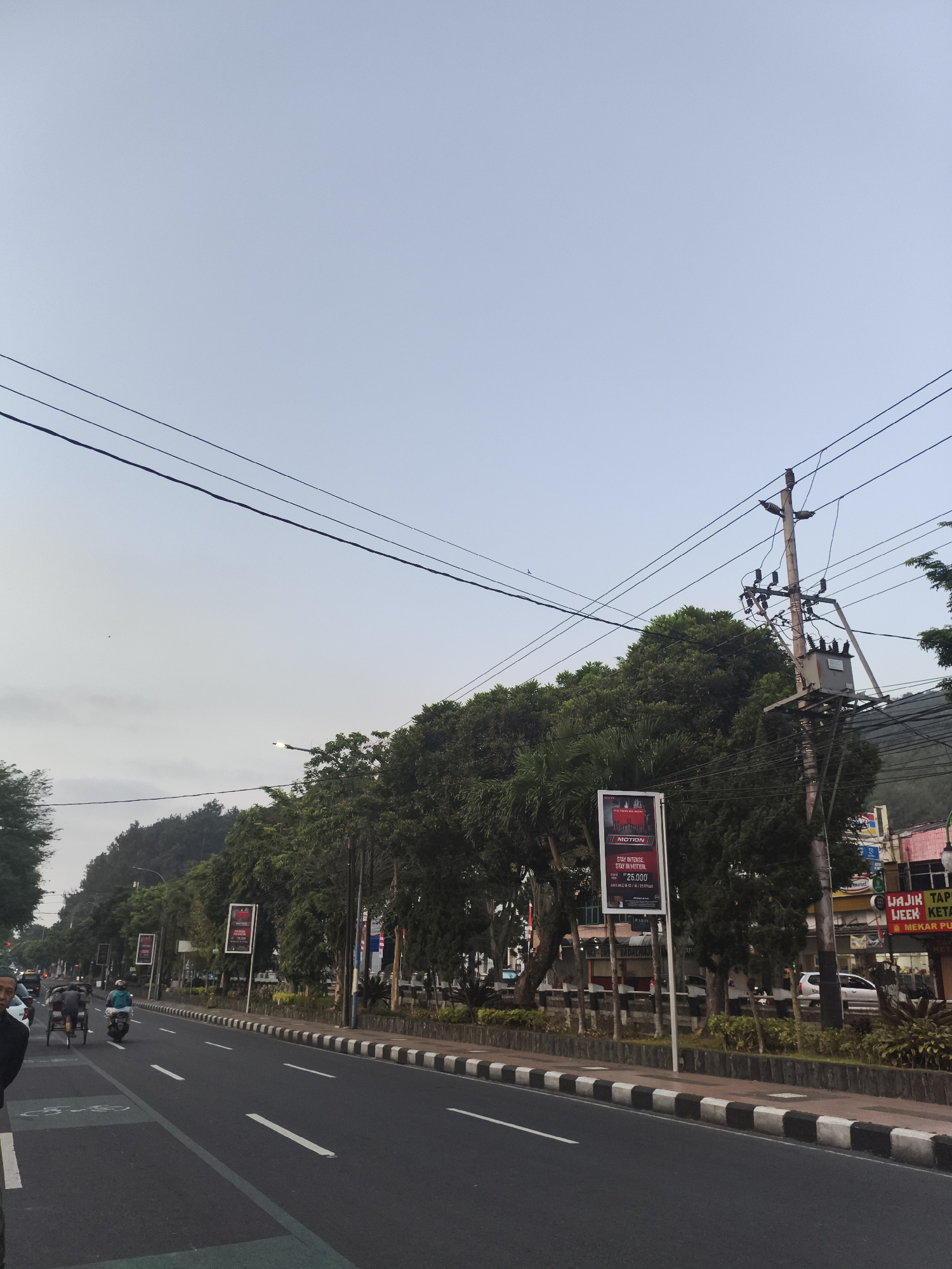The image depicts a street scene in Magelang, Indonesia, likely in the early morning or late afternoon given the soft light. A paved road with multiple lanes is visible, bordered by a sidewalk with a black and white striped curb. On the left side of the road, there are a few people on bicycles and a motor vehicle. Further down the street, a row of trees lines the sidewalk, behind which are buildings and what appears to be a hillside covered in vegetation. Overhead, power lines crisscross the sky, supported by utility poles. One pole, prominently featured, has a transformer and insulators. Several billboards are placed along the roadside, advertising various products and services. The sky is overcast and pale blue.