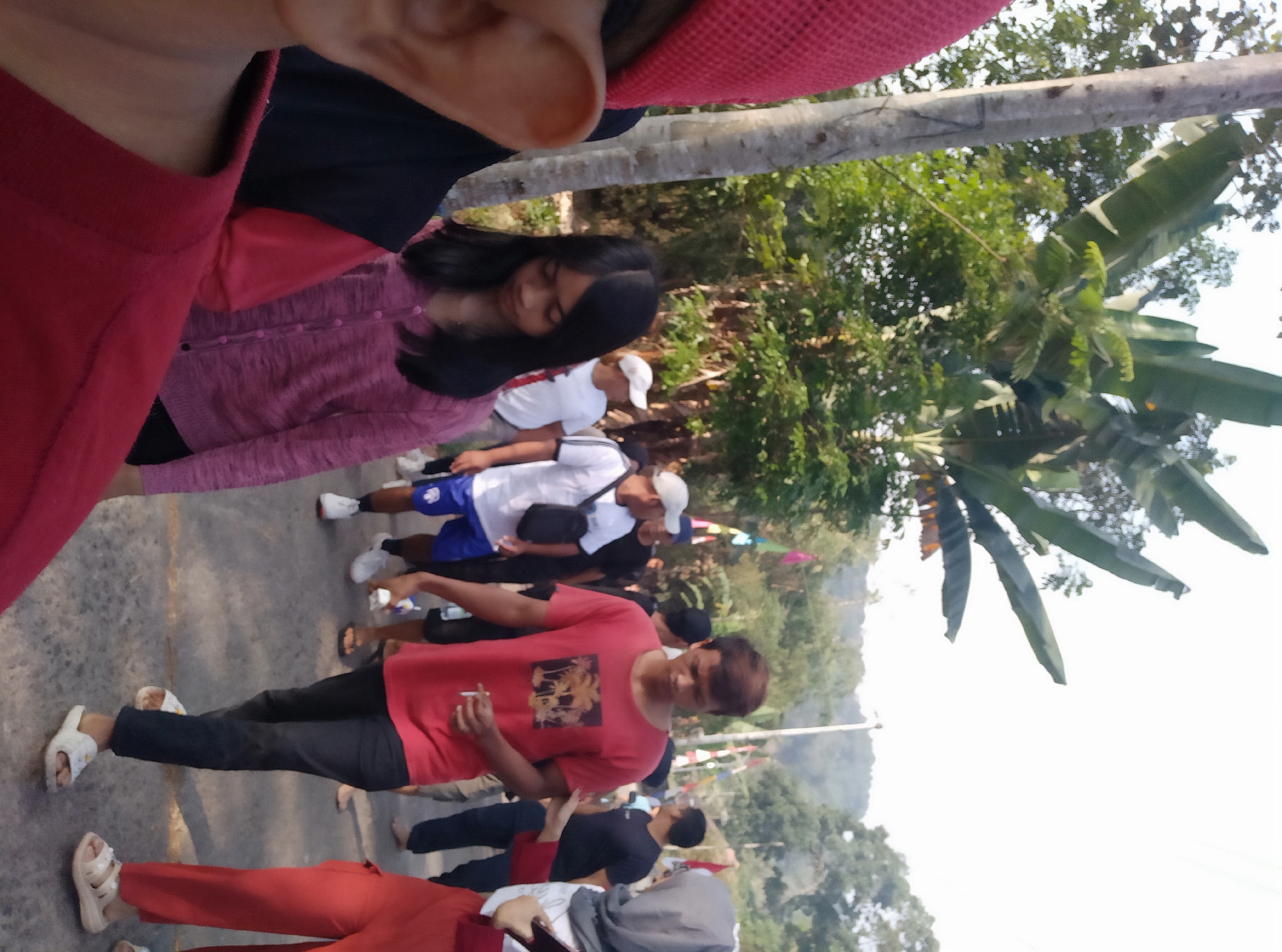 The image shows a group of people walking along a paved street in Lopait, Indonesia. The street is lined with trees, and a pole with banners is visible in the background. In the foreground, a person wearing a red hat and red shirt is partially visible on the left. To their right, a young woman with long black hair is wearing a pink cardigan. Further down the street, a man in a red t-shirt with a graphic print is walking, holding a cigarette. Other individuals, some wearing white shirts and blue shorts, are also present. The overall scene appears to be a daytime outdoor gathering.
