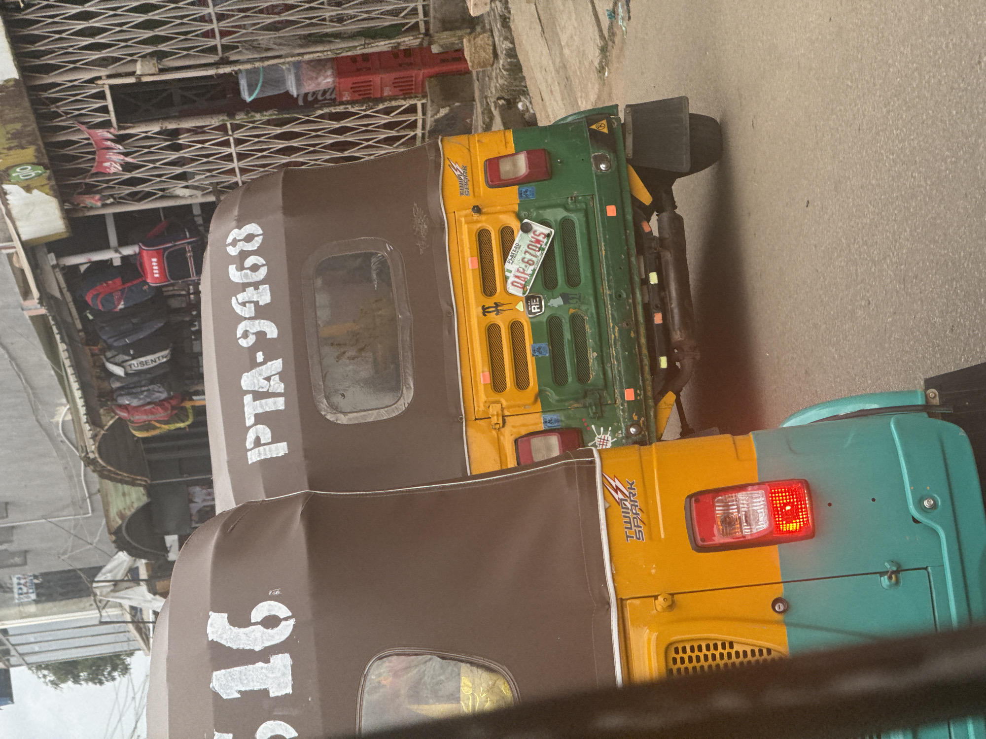 Two auto rickshaws are parked side-by-side on a street in Jos, Nigeria. The rickshaw on the left has "516" painted in white on its brown canvas top. The rickshaw on the right has "PTA-9468" painted in white on its brown canvas top and a license plate that reads "PLATEAU DAP-670WS". The rickshaw on the right is yellow and green, while the rickshaw on the left is yellow and teal. The rickshaws are parked in front of a building with a metal grate and shelves displaying bags and other merchandise. The street is paved and appears to be in a commercial area.
