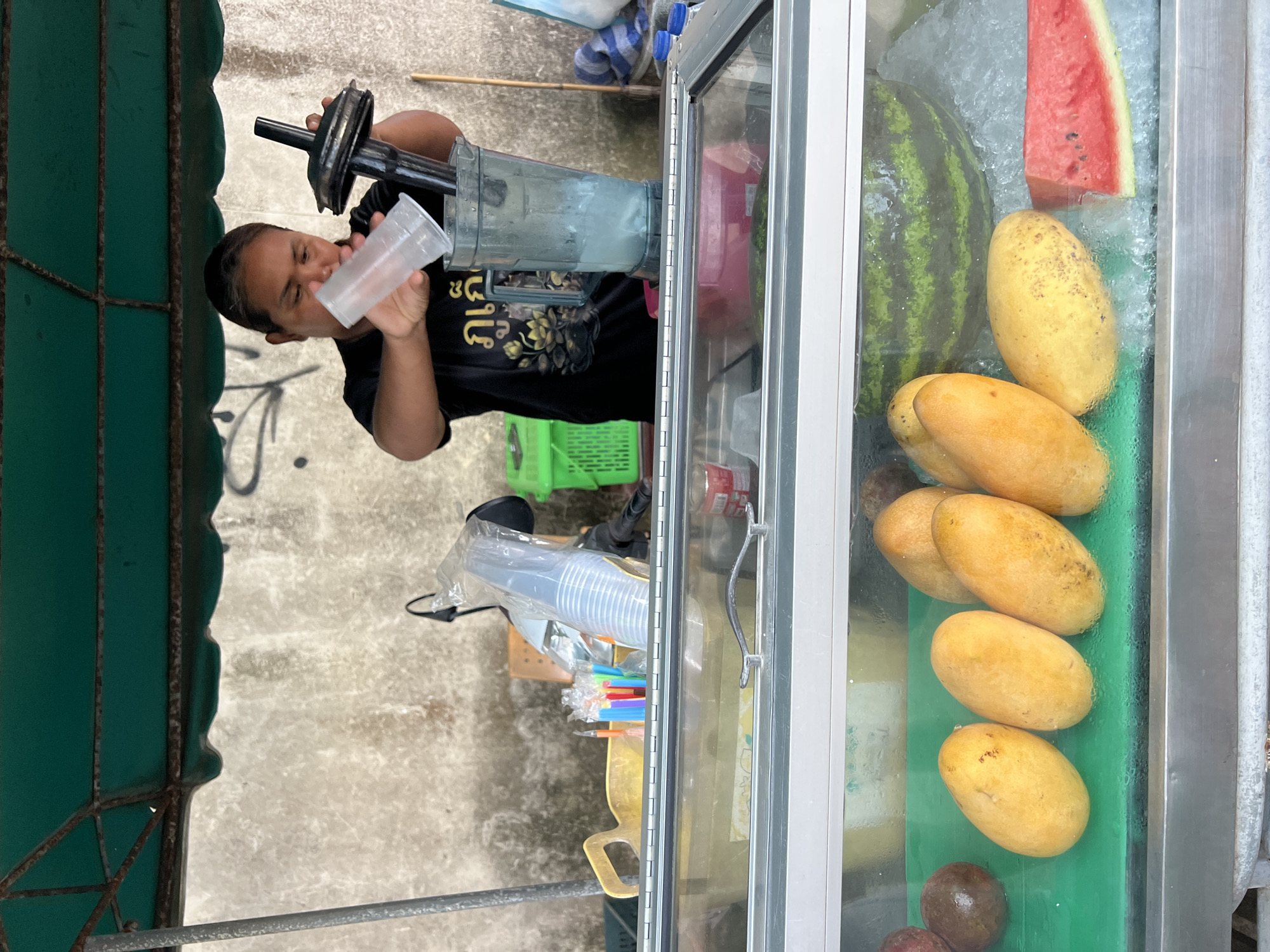 A woman is making a drink in a street stall. She is wearing a black t-shirt with a gold lotus design. She is pouring liquid from a clear plastic cup into a blender. The blender is on a counter in front of a display case filled with fruits. The fruits include mangoes, passion fruit, and watermelon. The street stall is covered by a green tarp, and a concrete wall is visible in the background.