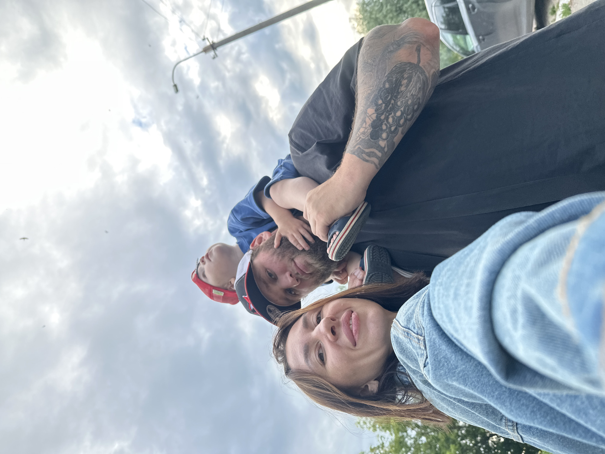 This media file captures a family in a selfie-style shot, seemingly outdoors during the day. The subjects include at least three people: an adult with extensive arm tattoos, another adult whose face is partially visible with their tongue sticking out, and a child wearing a red cap. The child is positioned on the back of the adult with tattoos. The scene includes a cloudy sky, a lamppost, and some greenery in the background, suggesting a park or an outdoor public space. The composition and angles indicate the photo was taken by one of the adults. The mood appears casual and playful, with the woman sticking out her tongue and the child being carried on someone's back. There is no visible text in the image. The mention of Votkinsk, Russia, provides a geographical context for the image.
