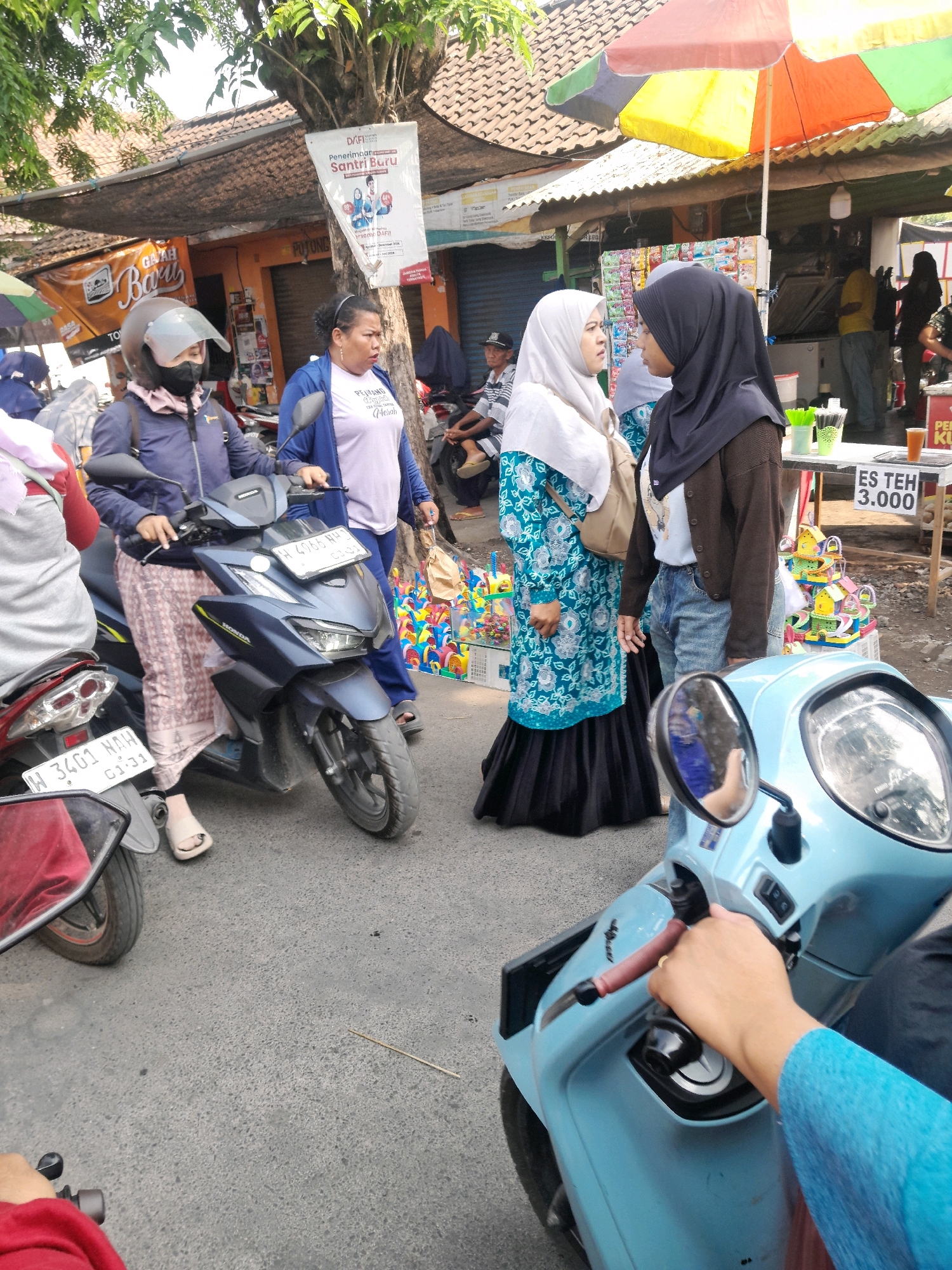 This is a medium-angle, eye-level shot of a street scene in Jogosatru, Indonesia, likely during the daytime, given the bright lighting. Three women are the primary subjects. The woman on the left is wearing a dark blue jacket, light pink headscarf, and a patterned skirt, seated on a dark blue motorcycle. She is wearing a helmet and a face mask. To her right, a woman in a light purple t-shirt and blue cardigan is standing, holding a brown paper bag. To her right, a woman wearing a white hijab and a blue and white patterned dress with a black skirt is standing, with a light brown backpack on her shoulder. Behind the women, there are several stalls and small shops, suggesting a market or roadside vendors. One stall has a sign that says "ES TEH 3.000" and displays colorful toy houses. There are other motorcycles visible in the scene, indicating a busy area with vehicle traffic. The ground is paved asphalt. The overall impression is a casual, bustling outdoor environment in an Indonesian city.