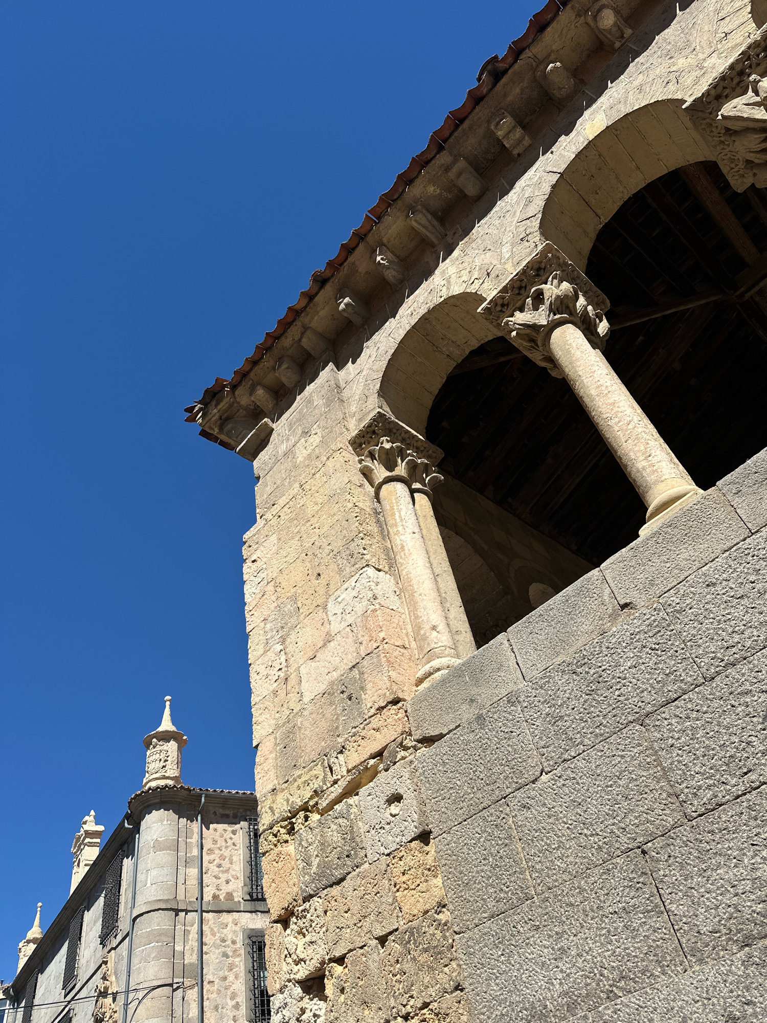 In La Lastrilla, Spain, a low-angle, close-up shot captures the architectural details of a stone building against a vibrant, clear blue sky. The weathered, light-colored stone of the building is visible, with some sections appearing rough and others smoothed. The structure features arched openings supported by columns with intricate, carved capitals. A section of a tiled roof is also visible at the top. To the left, another part of a building is partially visible, showing a tower with a decorative finial and barred windows. The overall scene is bathed in bright sunlight, casting distinct shadows and highlighting the textures of the stone. There are no people or animals present in the image.