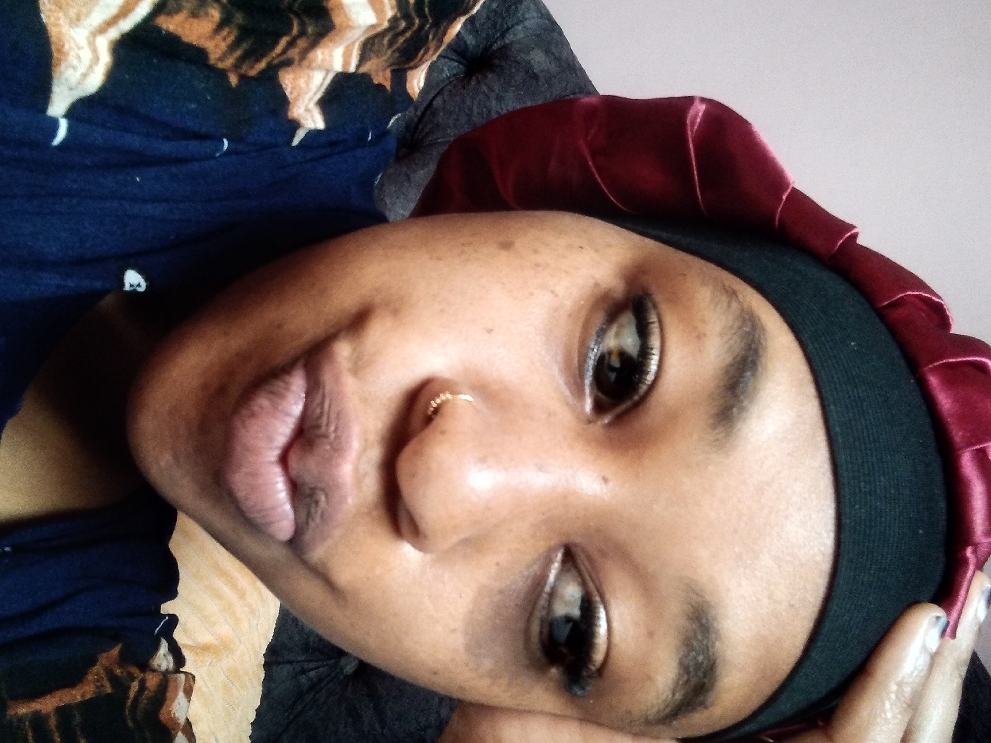 A close-up, medium-angle selfie shows a young Black woman wearing a black durag and a maroon satin bonnet. Her hand is resting on her forehead, and she is looking to her right with a pouting expression. She has a small gold hoop piercing in her left nostril and appears to be wearing subtle makeup, including eyeliner and mascara. The background is out of focus, but appears to be a soft, neutral color, possibly a wall. The woman is wearing a dark blue shirt with a pattern of orange and black shapes. The image was taken in Nyeri, Kenya.