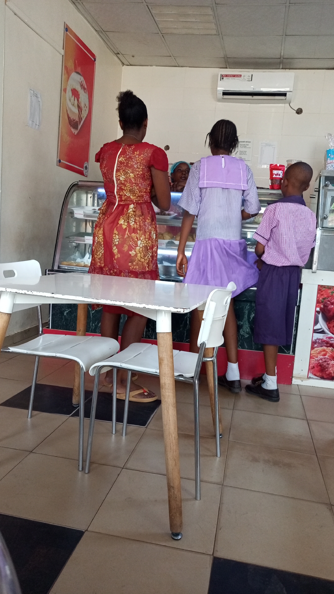 Three individuals stand at a display case inside a building, likely a bakery or fast-food restaurant. One is an adult woman wearing a red floral dress, and two are children in school uniforms with purple bottoms and striped tops. The adult is on the left, one child in the center, and the other on the right. Behind the display case, another adult is visible. In the foreground is a white table and chairs with wooden legs. The floor is tiled in alternating squares of brown and dark gray/black. A red poster is on the wall, and an air conditioner unit is mounted above the display case. This is in Narayi, Nigeria.