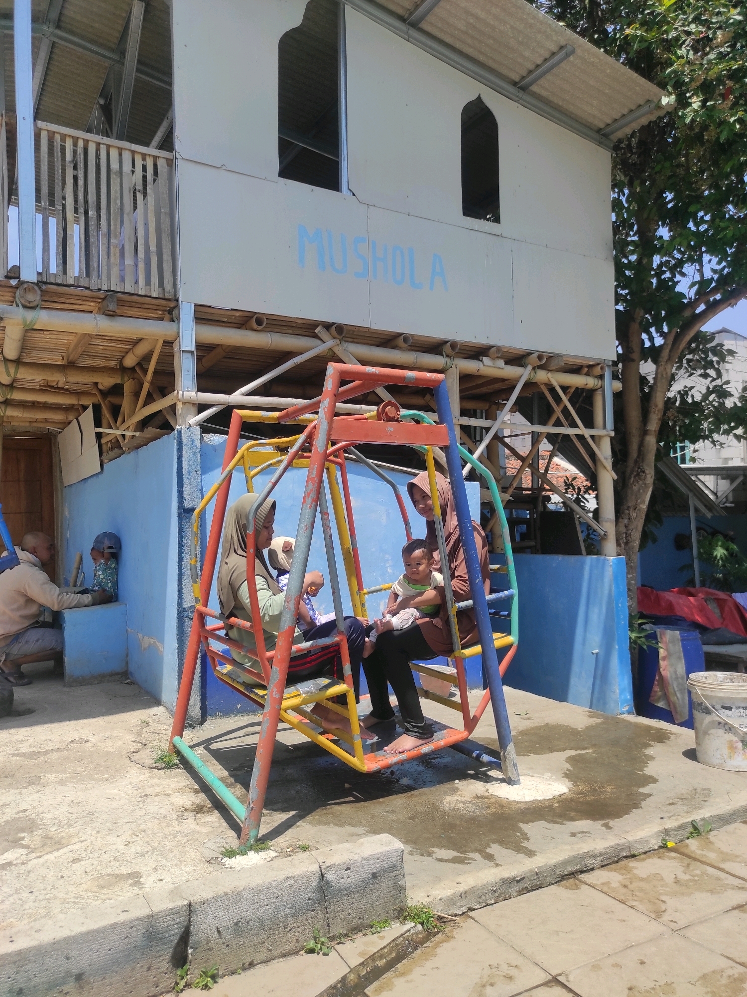 In Ngamprah, Indonesia, a multi-story building with a corrugated metal roof and pale blue siding, labeled "MUSHOALA," stands as a prominent structure. Below the building, a blue wall encloses a courtyard where a vibrant, multi-colored swing set is positioned. Three individuals are visible on the swing set. Two women, each wearing a hijab, are seated with a young child in between them. One woman is wearing a light green top and black pants, while the other wears a brown hijab and a black top. The child is dressed in a white and green outfit. Another individual, a man wearing a beige jacket and dark pants, is seen kneeling to the left of the swing set, interacting with a young child who is wearing a blue cap and a patterned shirt. The ground in the courtyard is paved with concrete, and a section near the swing set appears wet. Vegetation is present, with a tree visible to the right of the building and small plants growing along the edge of the paved area. A white bucket with some paint is visible to the right of the scene. The overall setting suggests an outdoor area associated with the "MUSHOALA" building.