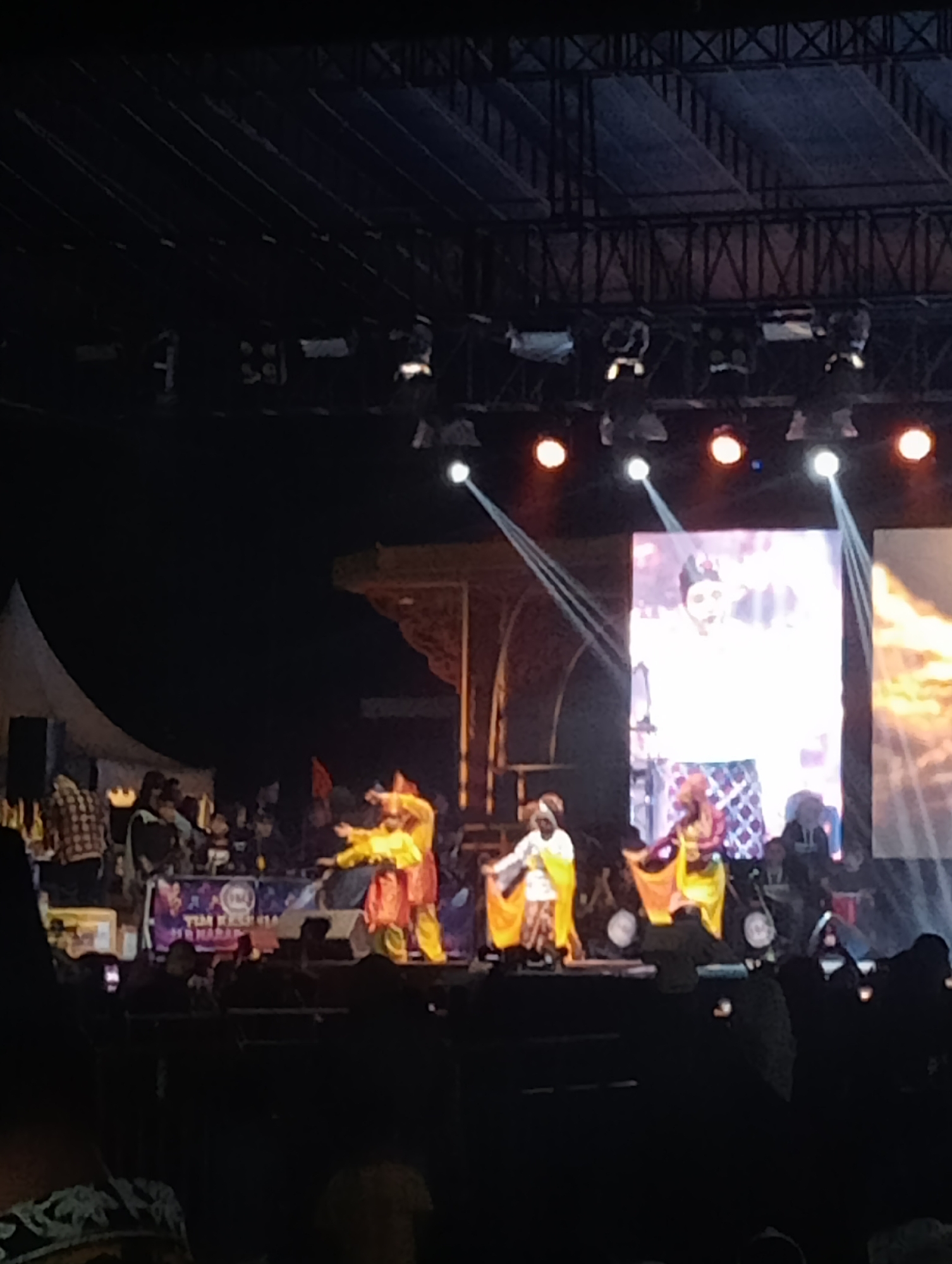 An outdoor night performance is taking place on a stage in Jambi, Indonesia. A large screen displays a person wearing a white garment and a dark head covering, with a patterned skirt. On the stage, performers in vibrant traditional costumes are dancing. Two performers in yellow and orange outfits are in the foreground, facing each other with outstretched arms, suggesting a synchronized movement. Another performer in a white and yellow outfit is on the left, with a yellow cloth draped over their arm. Spotlights illuminate the stage, creating beams of light that cut through the darkness. The audience is visible in the foreground, with silhouettes of people seated in rows, looking towards the stage. The stage is set against a dark background, with some structural elements visible above, including lighting rigs. The overall atmosphere appears to be festive and engaging.