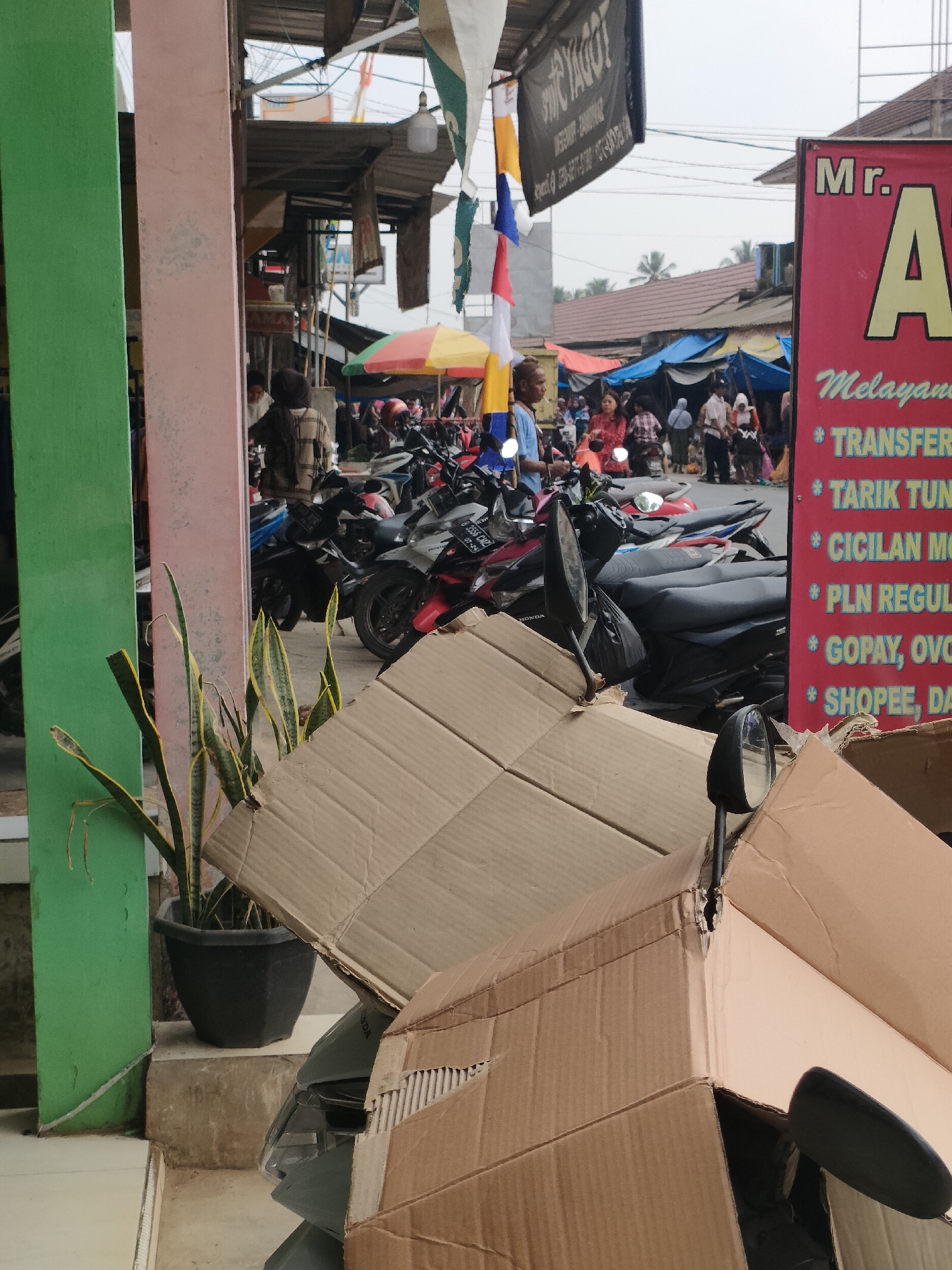 The image depicts an outdoor scene in Sukoyoso, Indonesia, likely a marketplace or street. In the foreground, several large cardboard boxes are piled up, obscuring parts of the view. A potted snake plant with green and yellow striped leaves is visible to the left, near a building with green and pink pillars. Behind the boxes, a row of parked motorcycles is evident. In the background, a busy street is filled with people and more motorcycles. Various stalls with colorful awnings and tarps suggest a commercial area. A prominent red sign with white and yellow text, reading "Mr. A Melayani TRANSFER... TARIK TUNAI... CICILAN MOTOR... PLN REGULER... GOPAY, OVO, SHOPEE, DANA," is positioned on the right side of the frame, indicating a service provider. The sky is overcast.