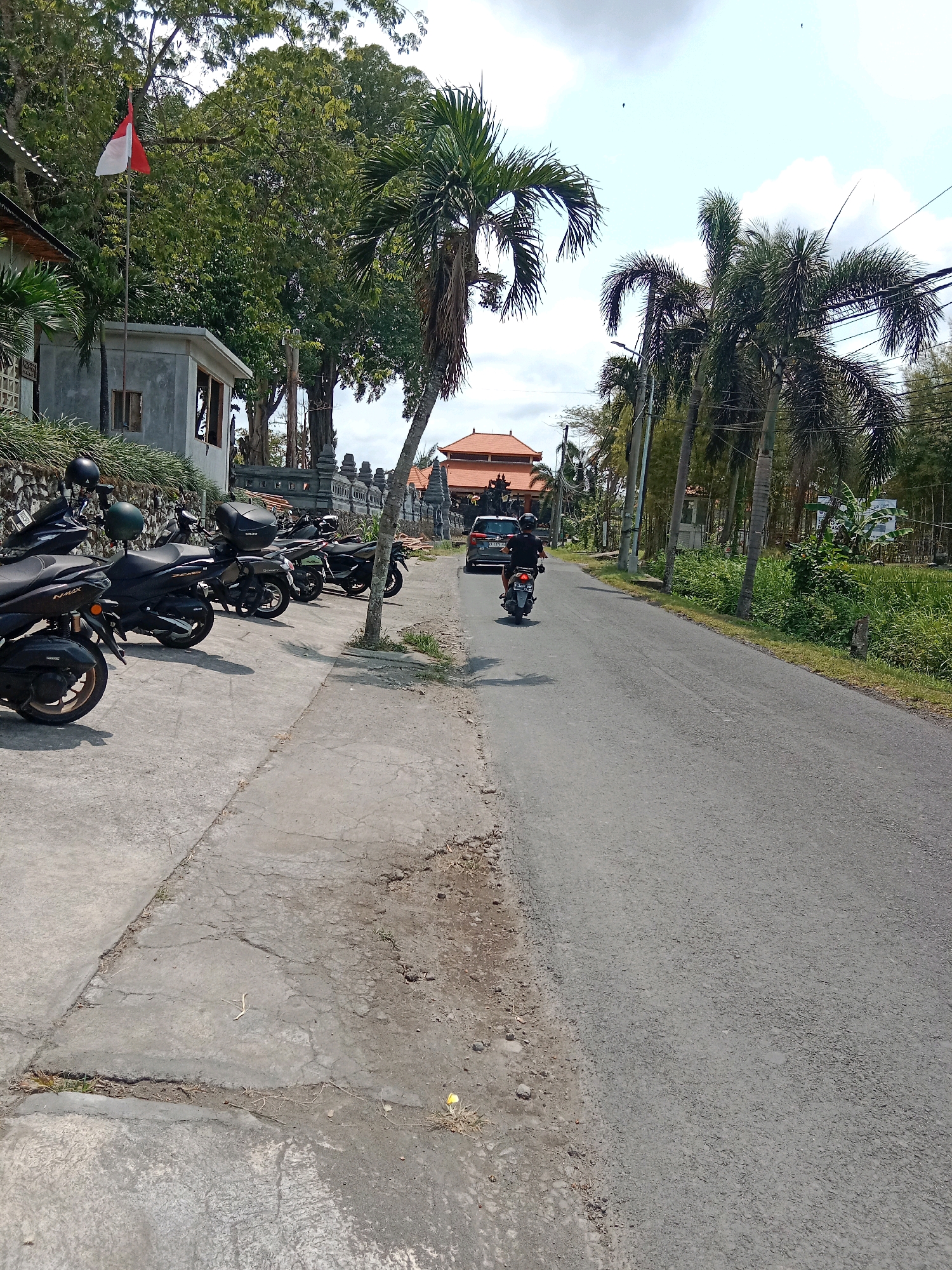 In the city of Mengwi, Indonesia, a paved road runs horizontally across the frame, bordered on the left by a concrete sidewalk where several parked motorcycles are lined up. The motorcycles are predominantly dark in color, with some featuring storage boxes on the rear. A red and white Indonesian flag flies on a flagpole near a low-rise building with a grey facade and a small window. Further down the road, a white car is visible, and a person on a motorcycle is riding away from the camera. The environment is characterized by lush green trees and vegetation, with palm trees lining the right side of the road. The sky is partly cloudy. The scene appears to be a street in a tropical region, possibly a residential or commercial area, given the presence of parked vehicles and buildings.