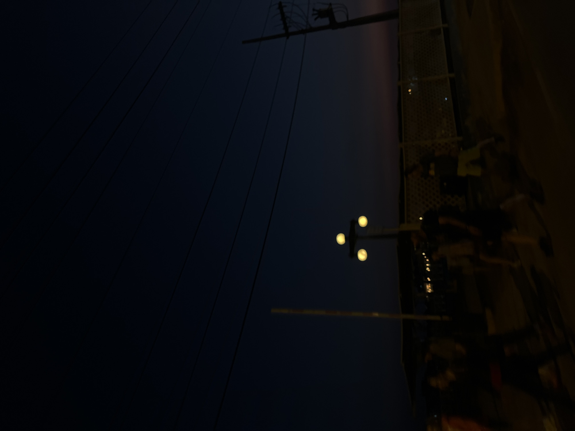 In Miraflores, Peru, at dusk, several individuals are walking along a pathway. The sky is a deep, dark blue, with several visible power lines stretching across the upper portion of the frame. A street lamp with three glowing orbs is positioned in the mid-ground, casting a warm light. To the right, a utility pole with insulators is visible. A light-colored fence runs horizontally in the background. The figures are blurred due to motion, suggesting movement and activity. The overall scene depicts a public space as evening transitions into night.
