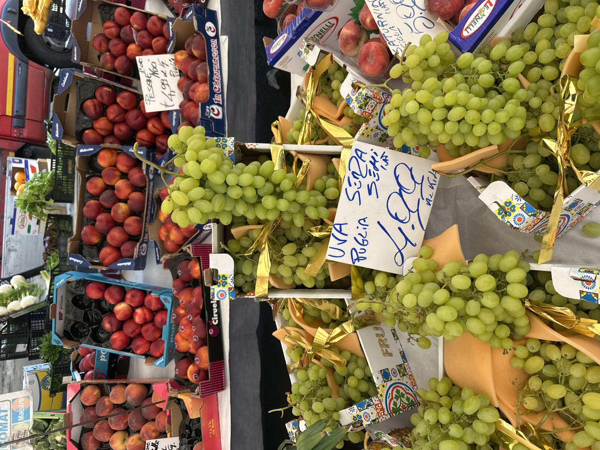 The image depicts a vibrant fruit stall, likely in Milan, Italy, overflowing with fresh produce. In the foreground, large bunches of green grapes are prominently displayed in white boxes, some adorned with golden ribbons. A price tag with handwritten Italian text, "UVA SERA SEMI 1,00," indicates the sale of seedless grapes for 1.00 unit of currency per portion. Behind the grapes, boxes of ripe peaches and nectarines are arranged. To the left, a sign with "Maestro" is visible. In the background, more crates of fruit and vegetables can be seen, along with glimpses of what appears to be a red vehicle, possibly part of a market stall setup. The overall scene suggests a bustling outdoor market environment.