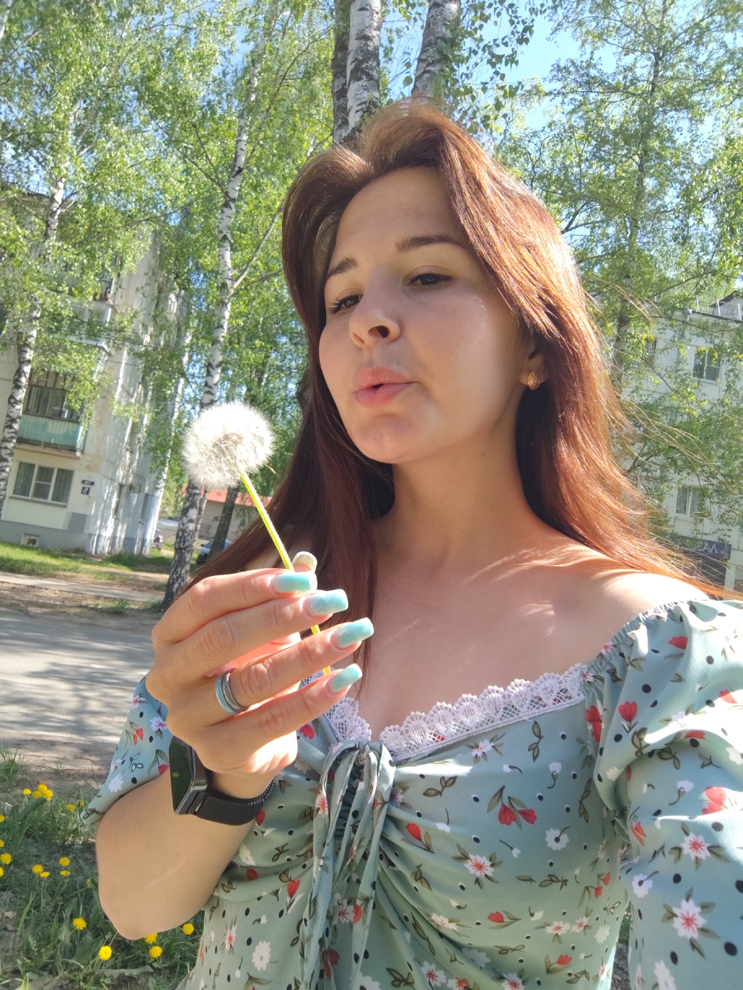 A young woman with medium-length reddish-brown hair is captured in a sunlit outdoor scene in Chaikovsky, Russia. She is shown in a mid-shot, looking towards the left, with her lips pursed as she blows on a white, fluffy dandelion held in her right hand. She has fair skin and is dressed in a light green, off-the-shoulder dress patterned with small red and white flowers, green leaves, and black dots, accented with white lace trim along the neckline.
Her right hand features long, light blue nails with white tips, and she wears a ring with blue and grey tones on her right ring finger. A black digital smartwatch with a mesh band is visible on her left wrist. Gold-colored earrings, possibly shaped like shells, adorn her ears.
The background is dominated by tall, slender birch trees with lush green leaves, indicative of spring or early summer. Behind the trees, light-colored apartment buildings with visible windows and balconies are present, suggesting an urban residential setting. A paved path or road runs across the mid-ground, and patches of bright yellow dandelions are visible in the foreground grass. The sky is clear and blue, signifying a bright, pleasant day. The image captures a serene and joyful moment.