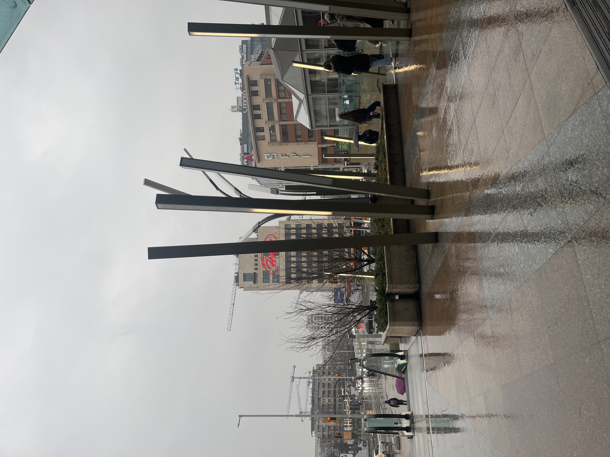 The image shows a rainy cityscape in Sofia, Bulgaria. Foreground: Wet tiled walkway with metal light structures. Further away are buildings, cranes, traffic lights, vehicles, and pedestrians. People are walking under umbrellas. The scene is overcast and appears to be taken from an elevated location. The building with the red logo “A” is prominent. In the middleground there is a building with signs that read: “Tarva” and “OFFICER”.