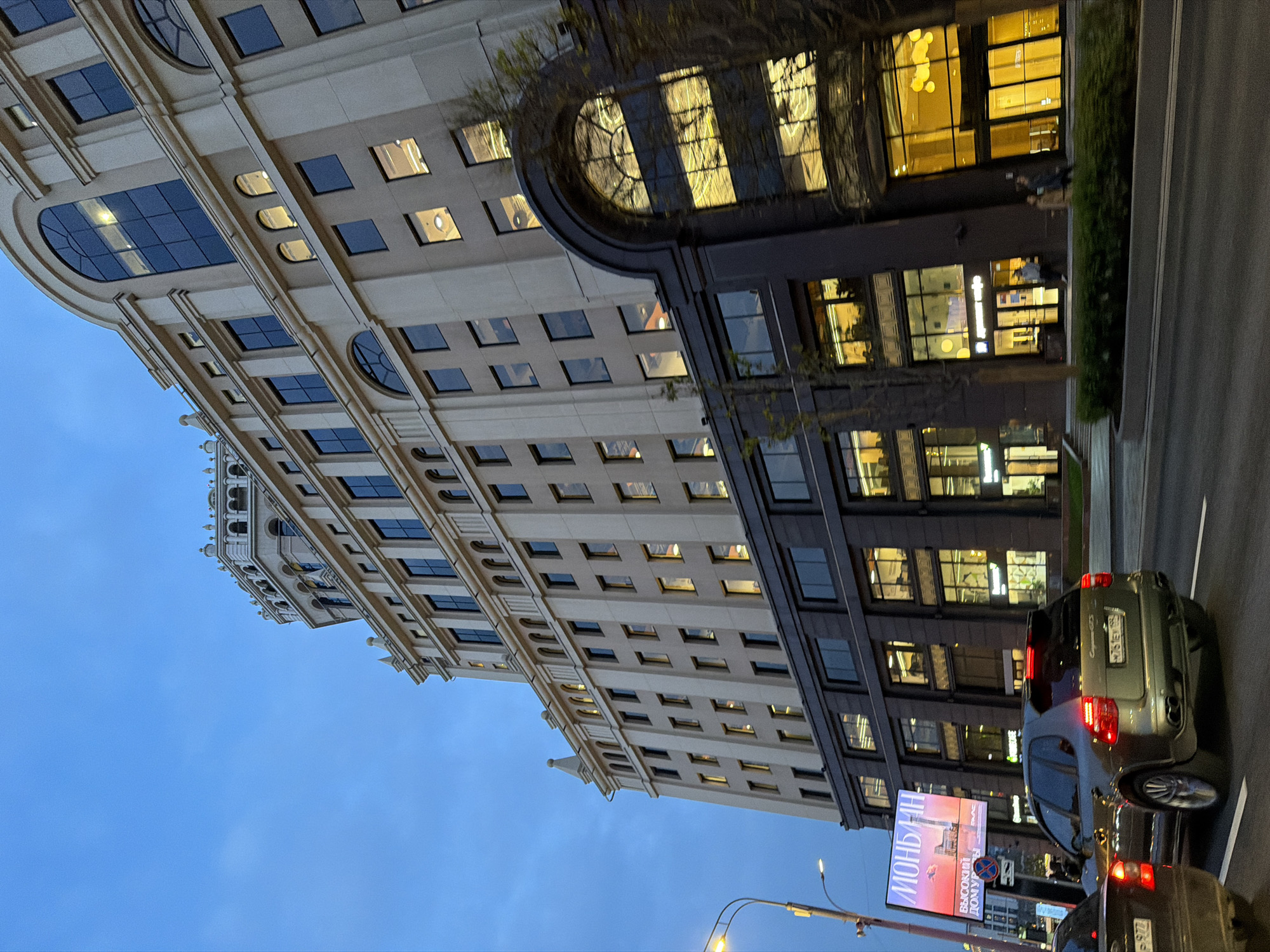 The image captures a street scene in Moscow, Russia at twilight. Two tall buildings dominate the view, one with a light beige facade and the other with a dark grey facade. Both buildings have multiple windows, with lights visible inside some. A street runs along the base of the buildings. Two cars are visible on the street: a green Porsche Cayenne and a dark-colored sedan behind the Porsche. An advertising display is also present near the cars. The sky above is a gradient of blue.