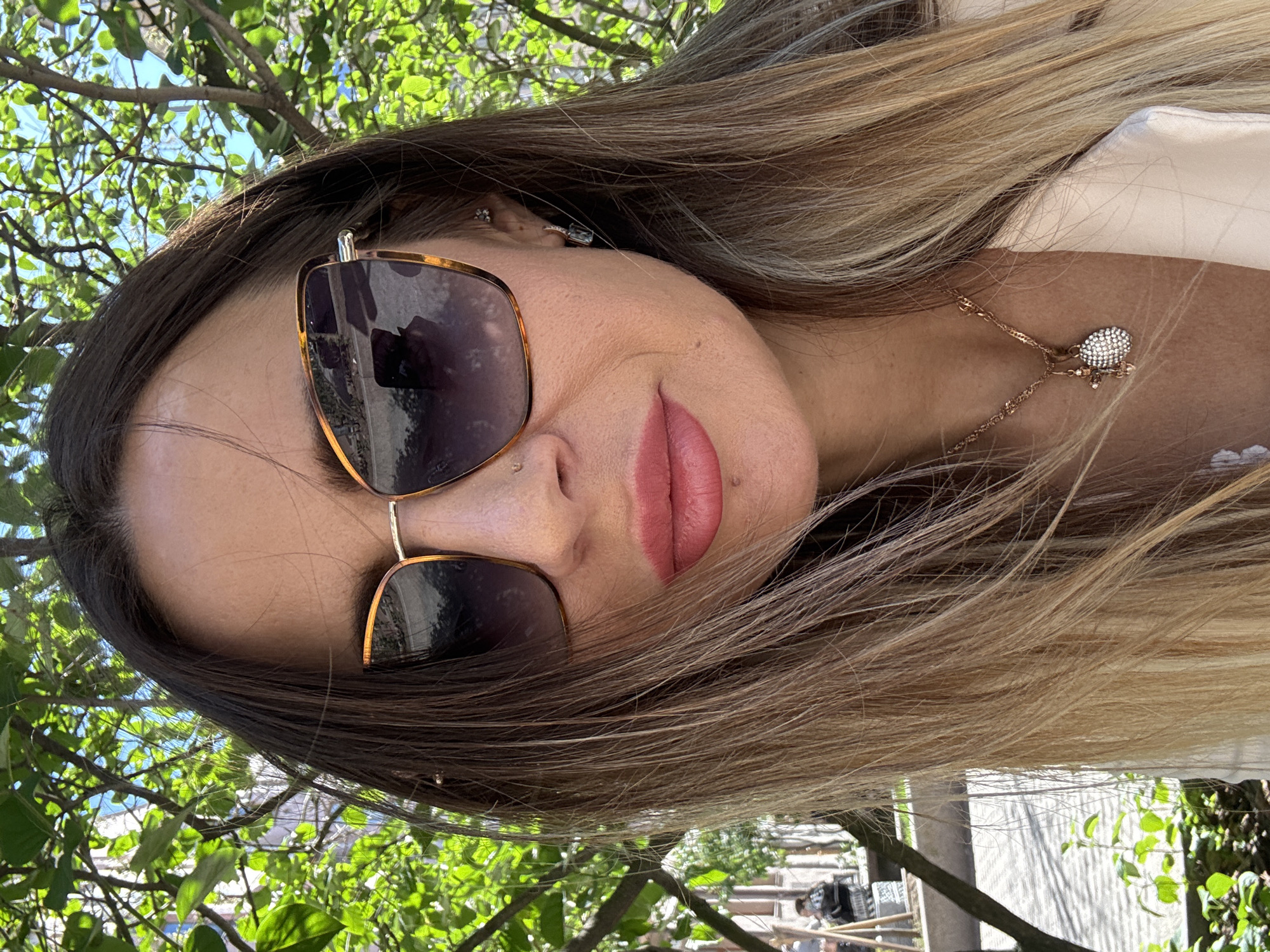 This media file is a portrait of a woman wearing oversized sunglasses with gold frames. She has long, light brown hair that falls around her face, and her lips are painted a soft pink. A delicate gold necklace with a small, diamond-encrusted pendant rests on her collarbone. She is wearing a light-colored, possibly silk, blouse.
The background suggests an outdoor setting with lush green foliage and dappled sunlight, implying it is daytime. The greenery appears to be from trees or bushes. The overall impression is one of casual elegance and a relaxed mood, possibly enjoyed on a sunny day. The rotation of the image suggests it may have been taken in a selfie orientation.
The visible elements do not provide specific cues about the exact location within Saint Petersburg, Russia, beyond the general impression of an outdoor, possibly urban or park-like environment. No text is visible in the image.