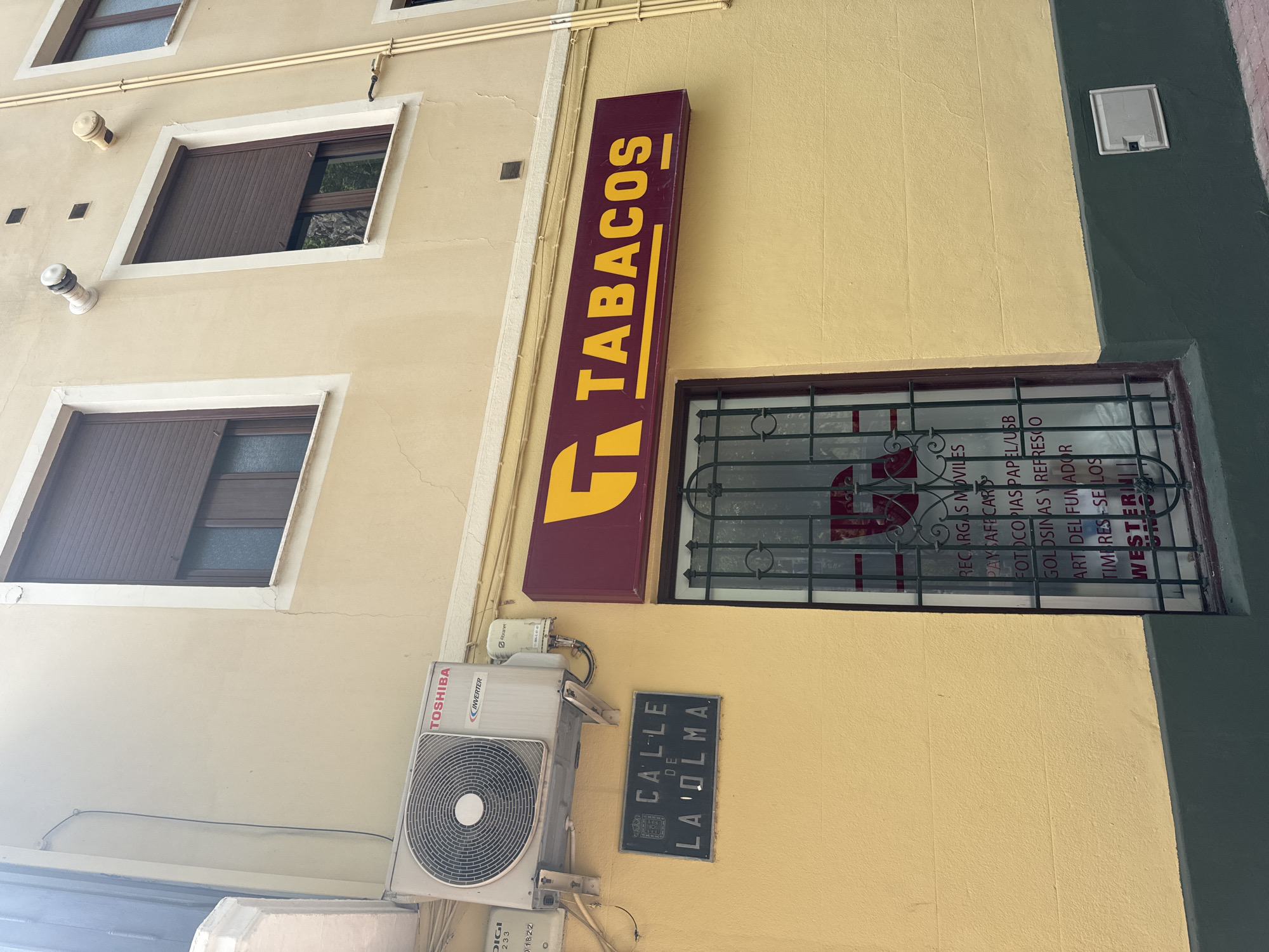 This is a vertically oriented, medium-angle shot of a building facade in Murcia, Spain. The building has a pale yellow exterior with dark green trim on the lower right. On the left side of the image, a white air conditioning unit is mounted on the wall. Above the air conditioner, a sign reads "TOSHIBA INVERTER". To the right of the air conditioner, a dark blue street sign displays "CALLE DE LA OLMA" in white lettering.
Further to the right, a large, vertical sign with a burgundy background and yellow text announces "TABACOS" in bold, sans-serif letters. Below this sign, a barred window reveals some of the interior. Text visible through the window includes "RECARGAS MOVILES", "PAYSAFECAR", "FOTOCO PIAS PAPEL/USB", "GOLDSINAS Y REFRESCO", "ART DEL FUMADOR", "TIMPRES SELLOS", and "WESTERN". The overall impression is of a shop or commercial establishment on a street in a European city. The lighting suggests it is daytime, and the weather appears clear. There are no people visible in the image.