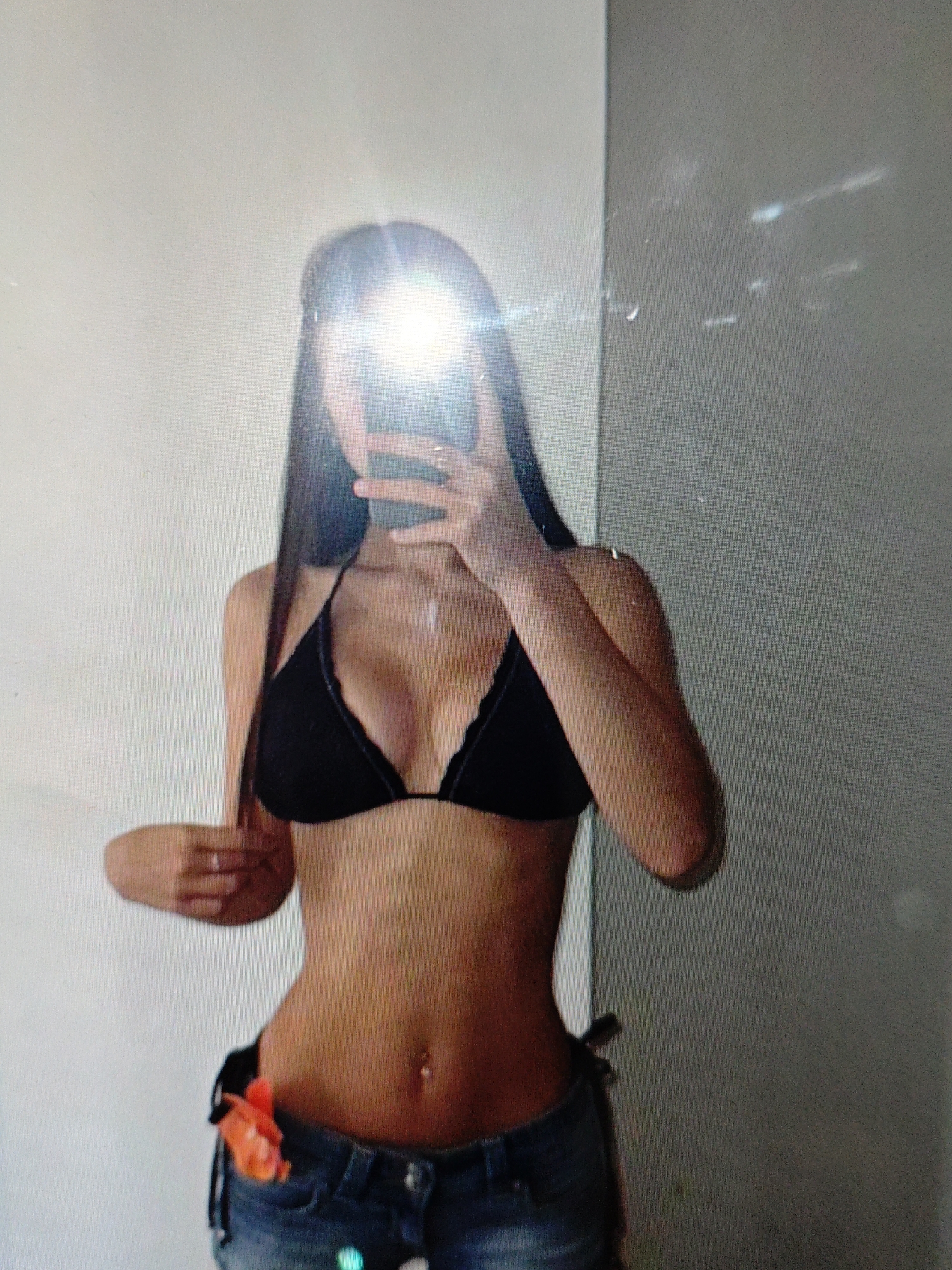 The image shows a person taking a selfie in a room. The person is wearing a black bikini top and blue denim shorts. They have long dark hair and a belly button piercing. There is an orange object tucked into the waistband of the shorts. The background consists of a white wall on the left and a grey wall on the right. The lighting in the room is bright, causing a lens flare in the center of the image, obscuring the person's face. The overall impression is casual and personal.
