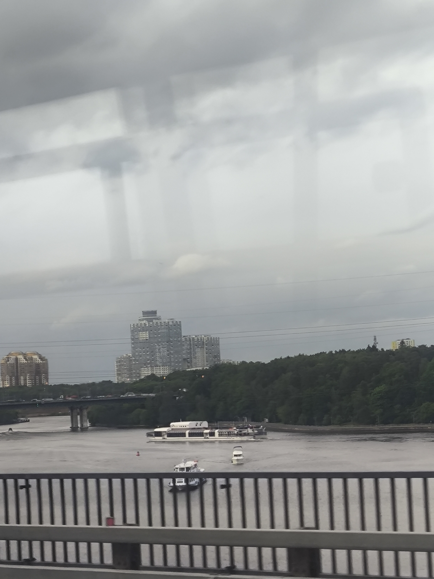 The image depicts a river scene, likely captured from a bridge. The sky is overcast with heavy clouds. In the distance, several buildings are visible, including a large, modern high-rise with a distinctive crown-like structure. A bridge spans the river in the mid-ground, with vehicles visible on it. Two boats are on the water. The foreground is dominated by the bridge's railing, which is composed of vertical bars. The overall scene evokes a sense of urban landscape with a focus on the river and its surroundings.