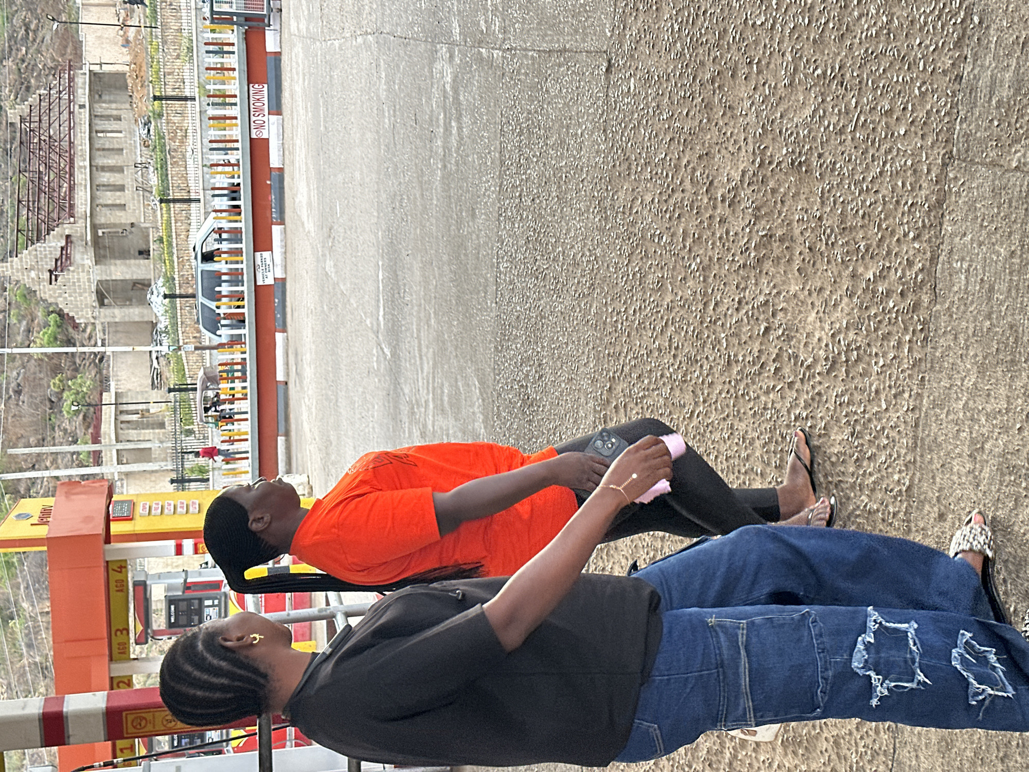 The image, rotated 90 degrees clockwise, captures two individuals standing at what appears to be a gas station or a service area under bright daylight, likely in Jos, Nigeria.
In the foreground, a person with braided hair, wearing a dark top (possibly a jacket or hoodie) and ripped blue jeans, is visible. This individual wears a distinctive gold earring and casual footwear. Slightly behind them, another person is seen wearing a vibrant orange t-shirt and dark trousers. This person also has styled dark hair and holds a light-colored object, possibly a phone or tissue, in their left hand. Both individuals appear to be looking upwards or forward, suggesting they are observing something or waiting.
The setting includes a paved ground area with a clear transition from a smoother texture to a rougher, pebble-like surface. To the left, several yellow and red gas pumps are visible, marked with numbers "1" through "4" and a partially visible "JENNY" logo. A red barrier with white stripes runs across the mid-ground. On this barrier, a clear "NO SMOKING" sign is displayed, along with a partially visible text indicating "VEHICLES ARE PARKED AT OWNERS RISK".
In the background, beyond a fence with colorful yellow, red, and white bars, unfinished concrete structures or buildings are visible amidst a rocky, hilly landscape. A white car can be seen parked behind the fence. The overall impression is an active, semi-urban environment.